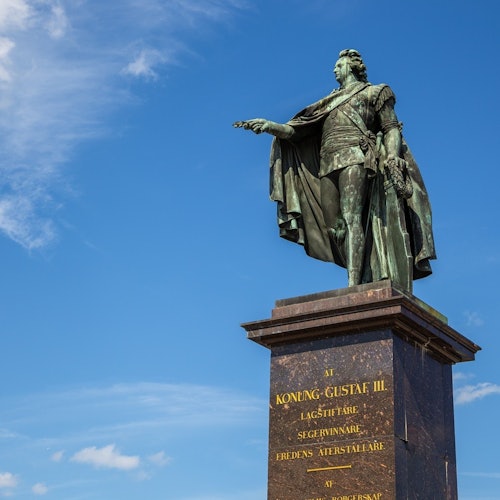 Bronsskulptur av en man i tidstypiska kläder på en hög sockel med ingraverad text mot en blå himmel med tunna moln.