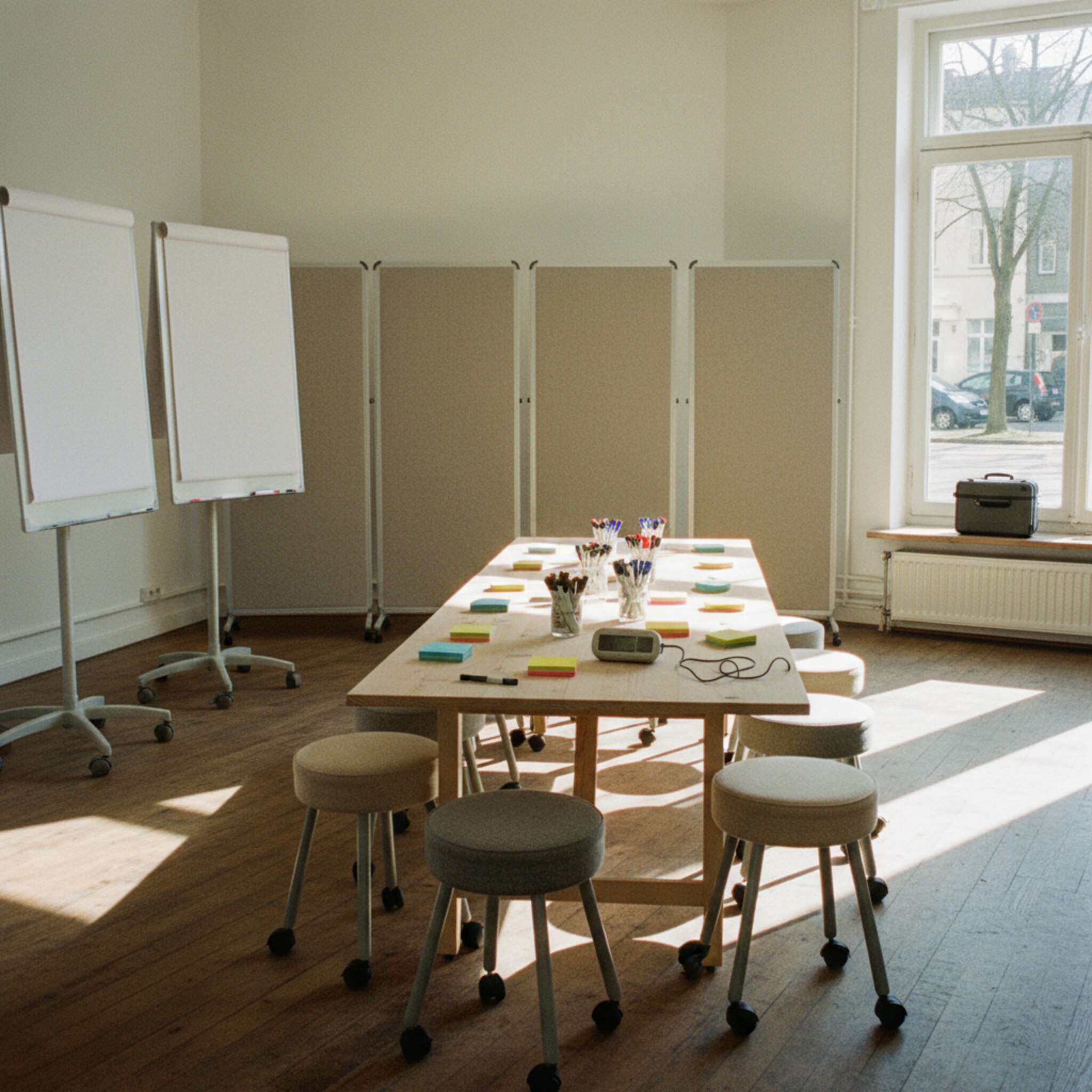 Ein Workshopraum mit beweglichen Whiteboards, Stapeln bunter Haftnotizen und rollbaren Hockern ist für eine Session vorbereitet. An der Wand stehen zusammengefaltete Stellwände, am Tisch liegen Marker und Timer. Durch ein Fenster fällt warmes Licht auf einen geparkten Moderationskoffer. Die Szene wirkt offen und startklar.