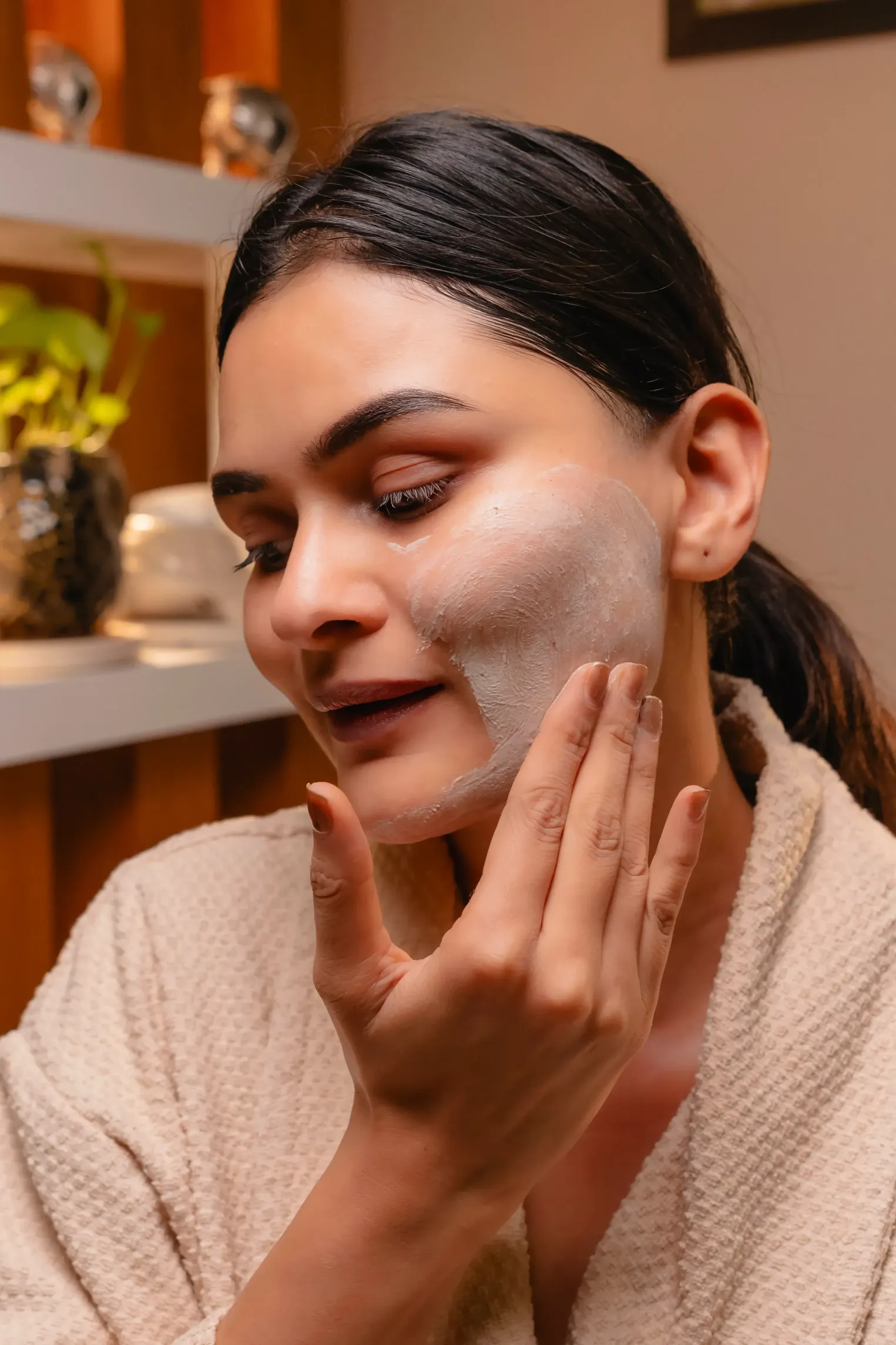 A woman applies a white face mask, smiling gently in a cozy, well-decorated environment.