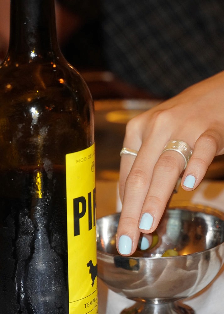 Close-up of a bottle of wine with a hand holding a metal cup, wearing silver rings.