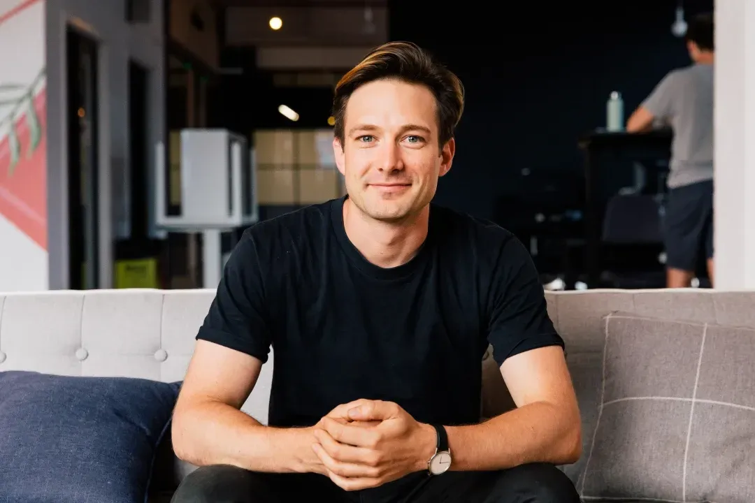 A person sits on a modern gray couch in a stylish, contemporary office setting, wearing a black t-shirt and watch, with hands clasped, surrounded by soft cushions and a blurred background of office furniture.