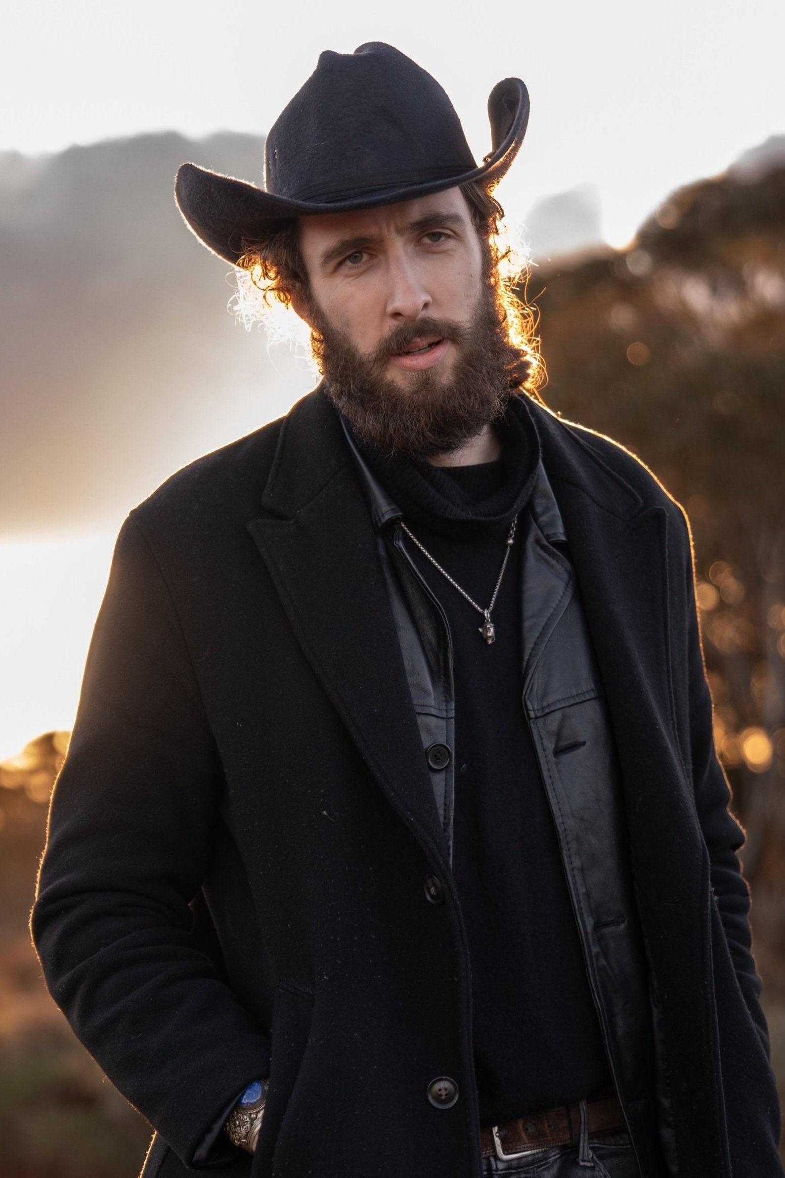 A man in a brown suit and cream silk scarf poses against a neutral background, exuding a composed and sophisticated demeanor.