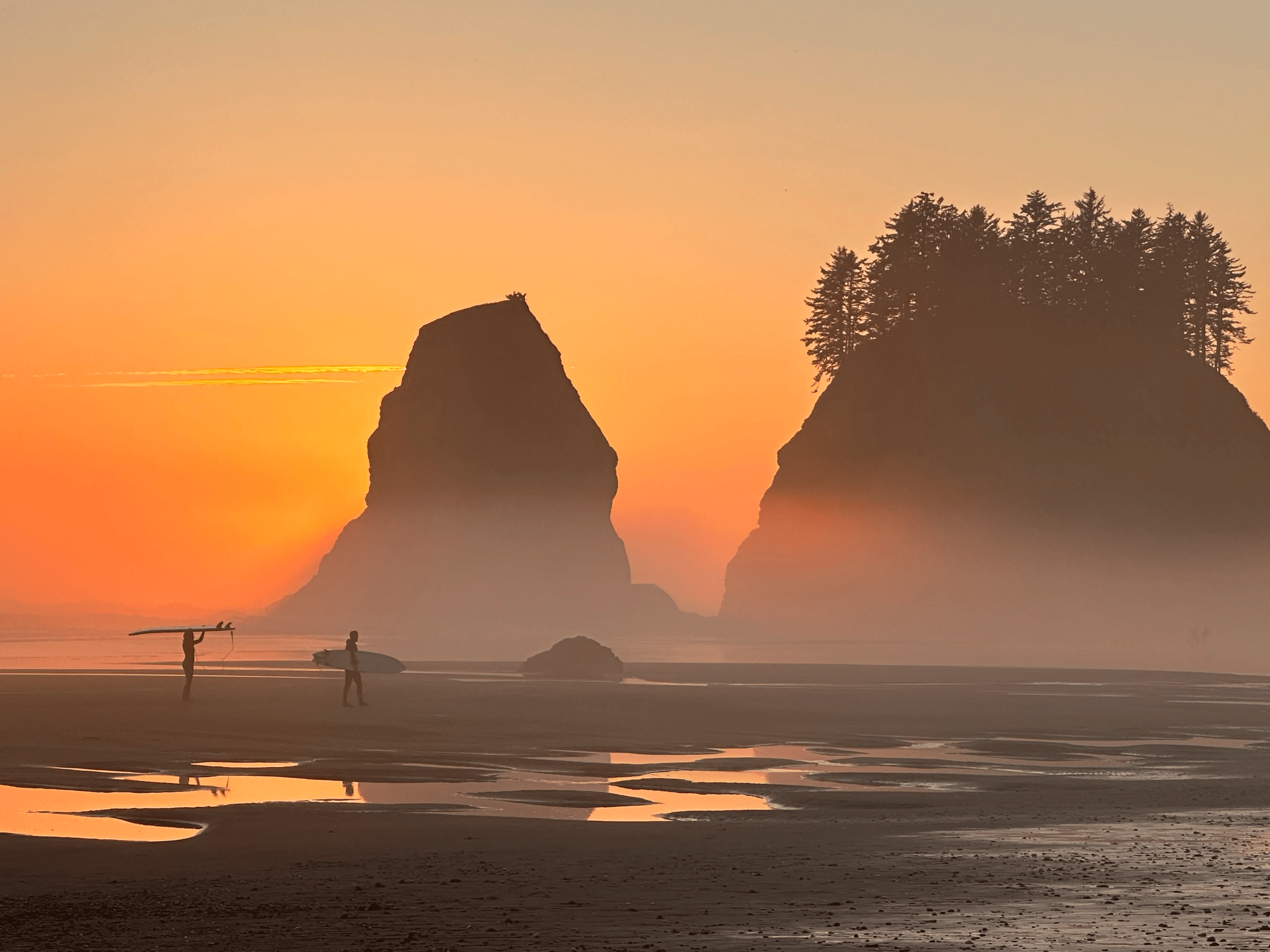 Two surfers carrying boards walk along a tidal beach silhouetted by sea stacks at sunset in Olympic National Park.