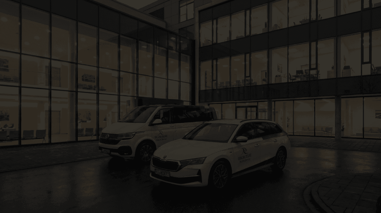 Zwei Rikscha Deluxe Fahrzeuge – ein VW Bus und ein Škoda Octavia – mit Beschriftung auf den Türen, geparkt vor einem modern beleuchteten Krankenhaus bei Nacht.​​​​​​​​​​​​​​​​