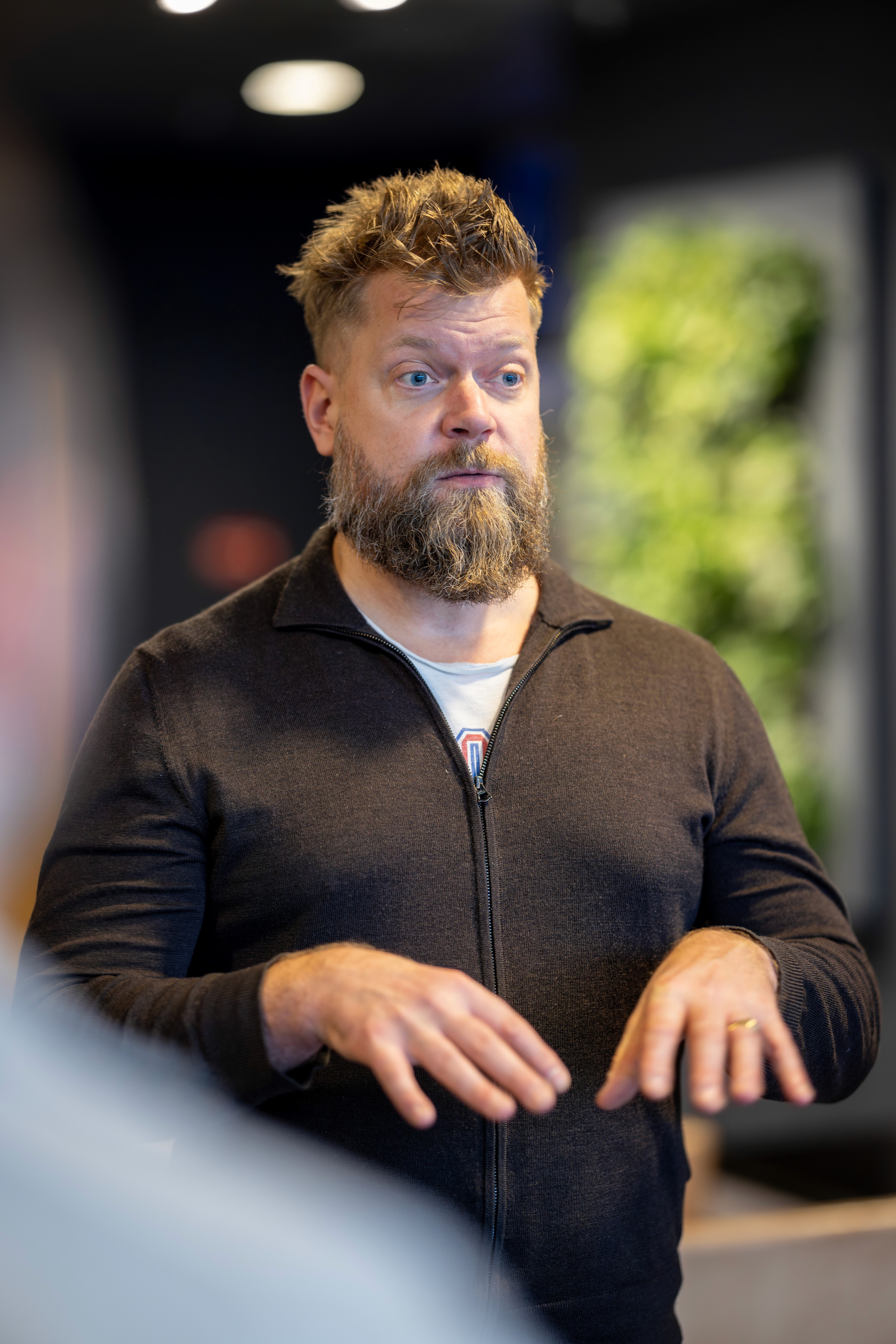 A man in a black shirt stands speaking, with a blurred figure in the foreground and a green background.