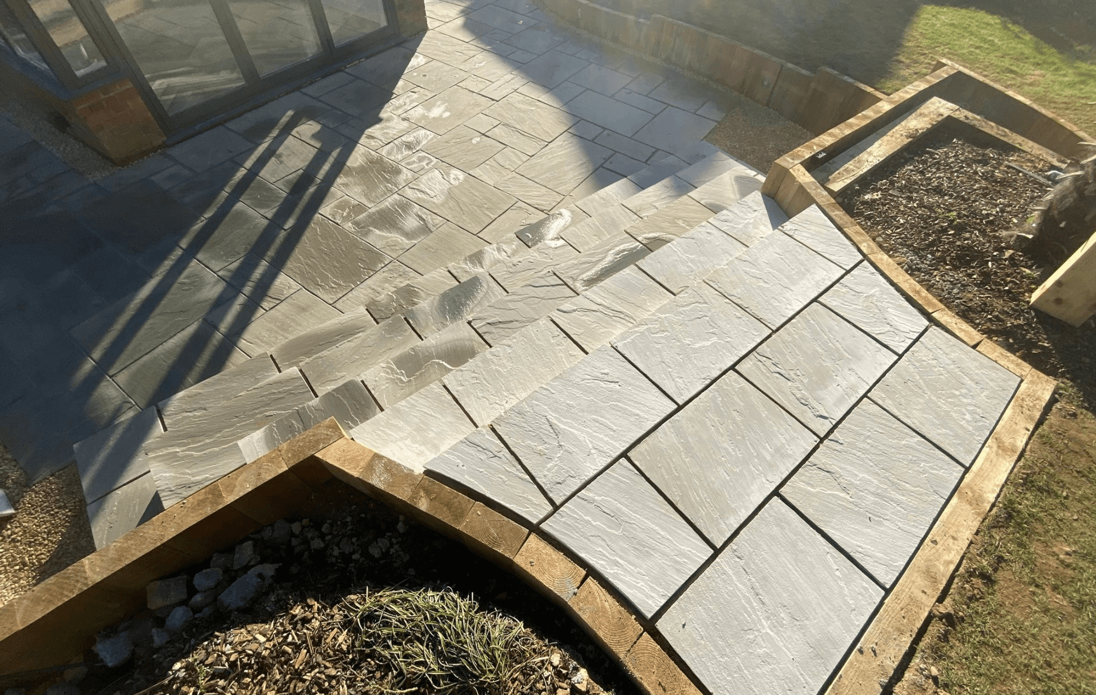 Multi-level Indian sandstone patio with bespoke steps, installed by PPM Builders in Northampton. High-quality paving with wooden sleeper edging for a stylish and functional outdoor space.