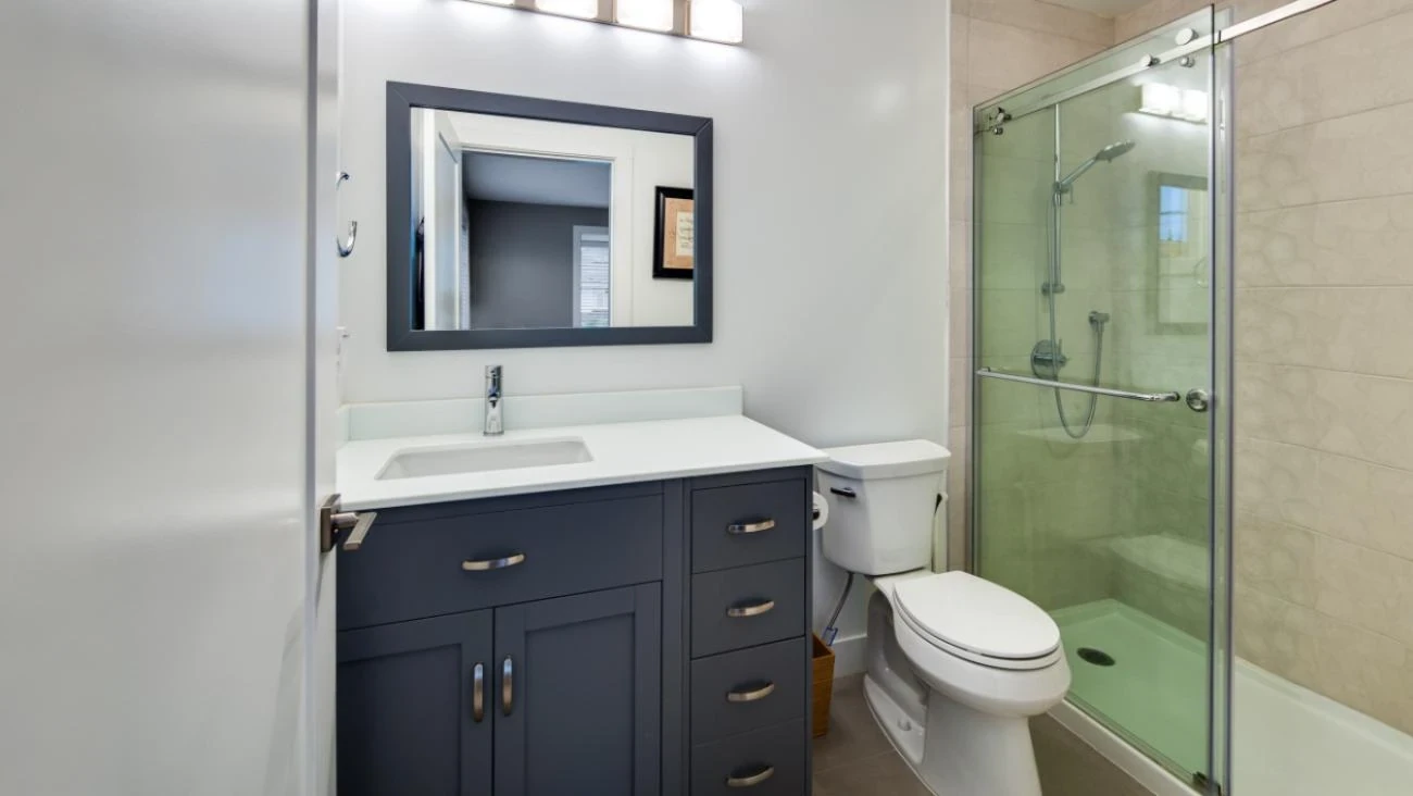 Modern full bathroom in Vancouver Custom Homes build with navy blue shaker-style vanity cabinet, white quartz countertop with undermount sink, black-framed mirror, frameless glass-enclosed walk-in shower, and contemporary neutral tile
