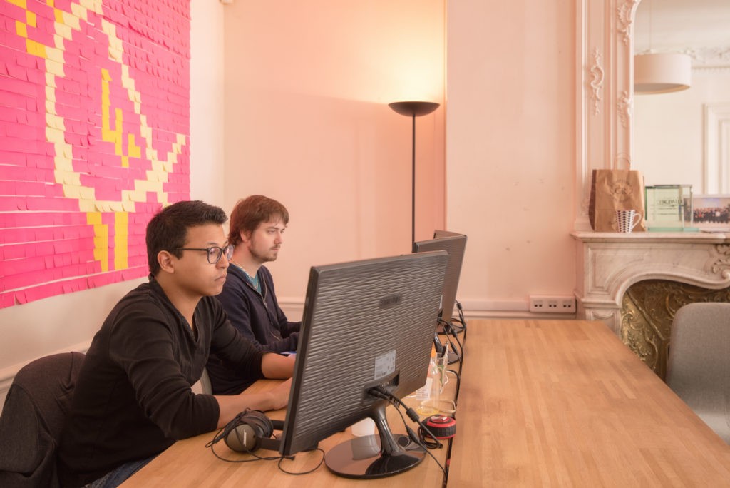Deux développeurs travaillent côte à côte devant leurs écrans, devant un mur de post-it roses