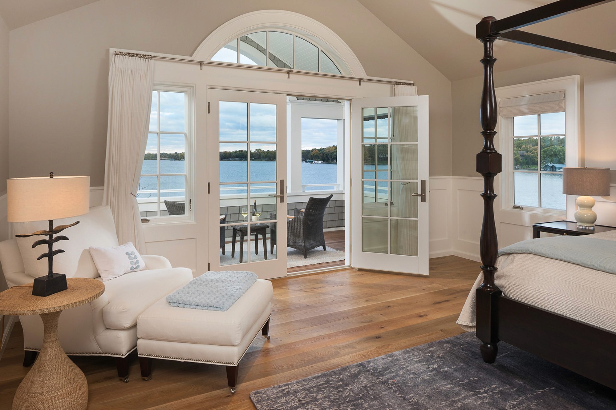 Coastal style master bedroom with a large four-poster bed and open French doors leading to a balcony with a lake view.