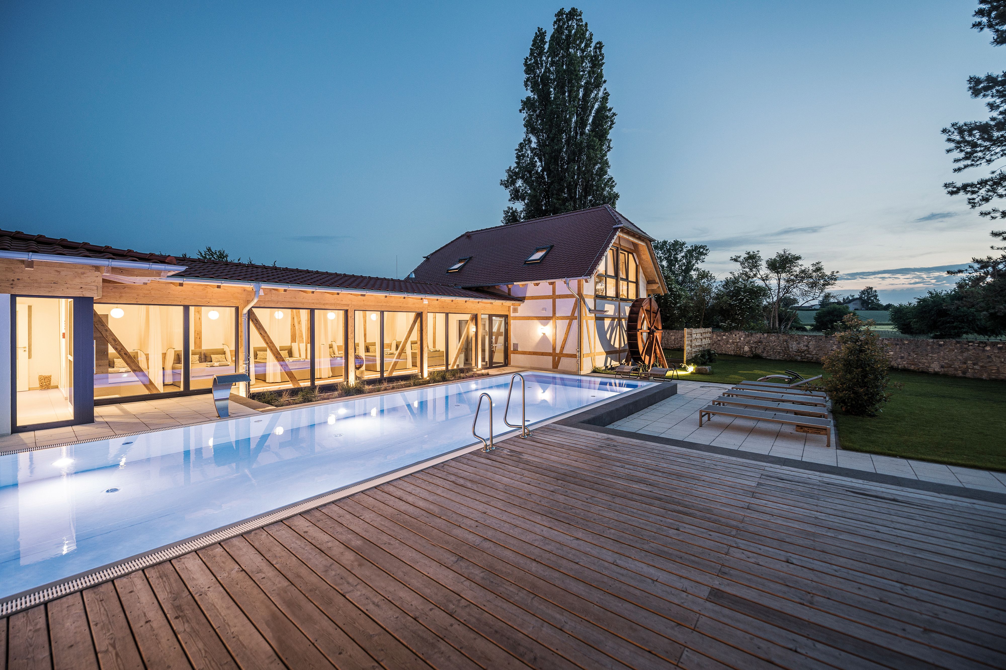 A luxurious outdoor swimming pool next to a modern, eco-friendly house with large glass windows and wooden accents, surrounded by trees and a serene landscape at dusk.
