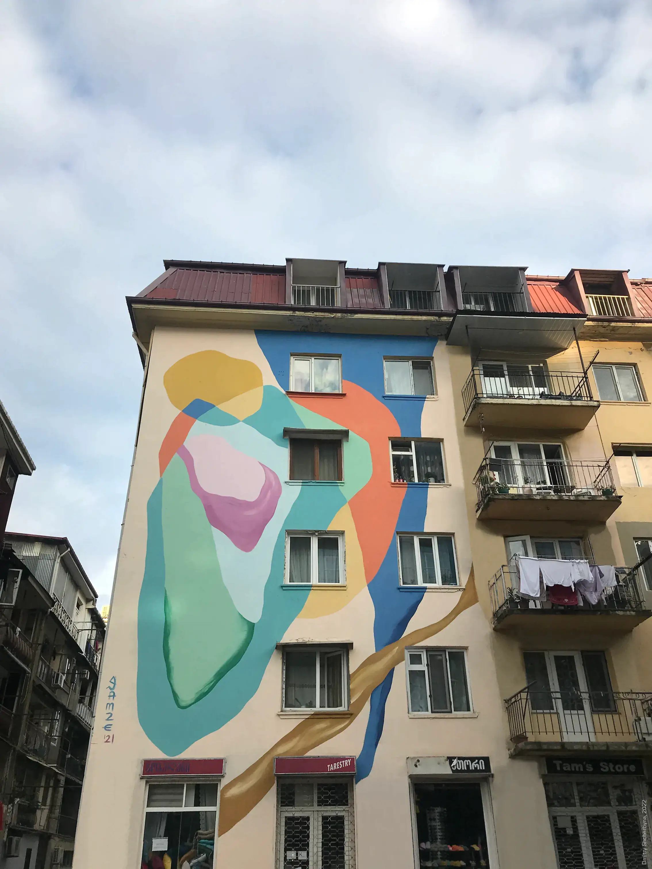 A mural on the side of an apartment building depicts two abstract figures in an embrace rendered in soft, overlapping shapes of blue, coral, mint green, yellow and pink.