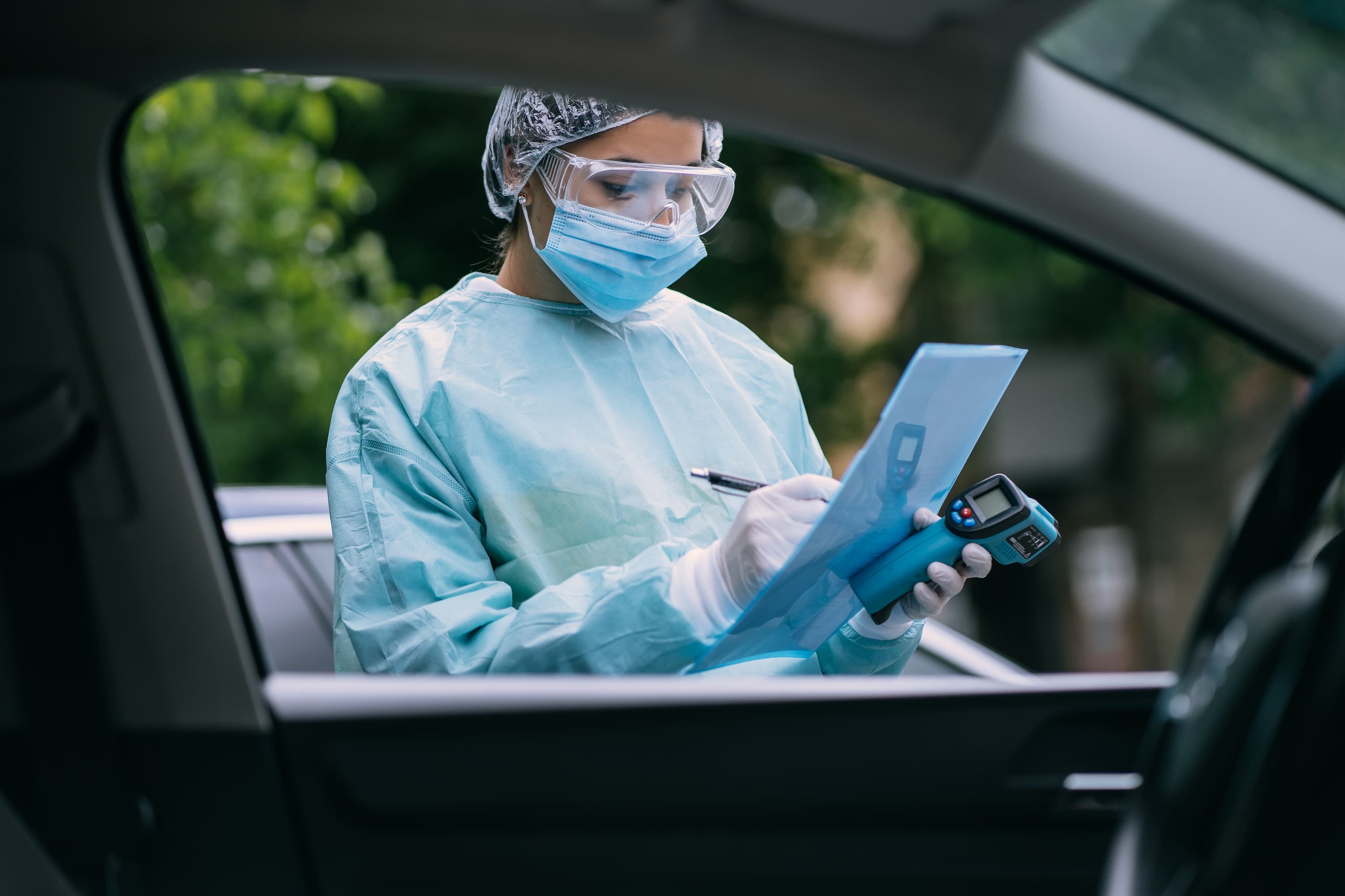 Healthcare worker in protective gear, including a mask, goggles, and gloves, recording information on a clipboard while holding a digital thermometer, viewed through a car window.