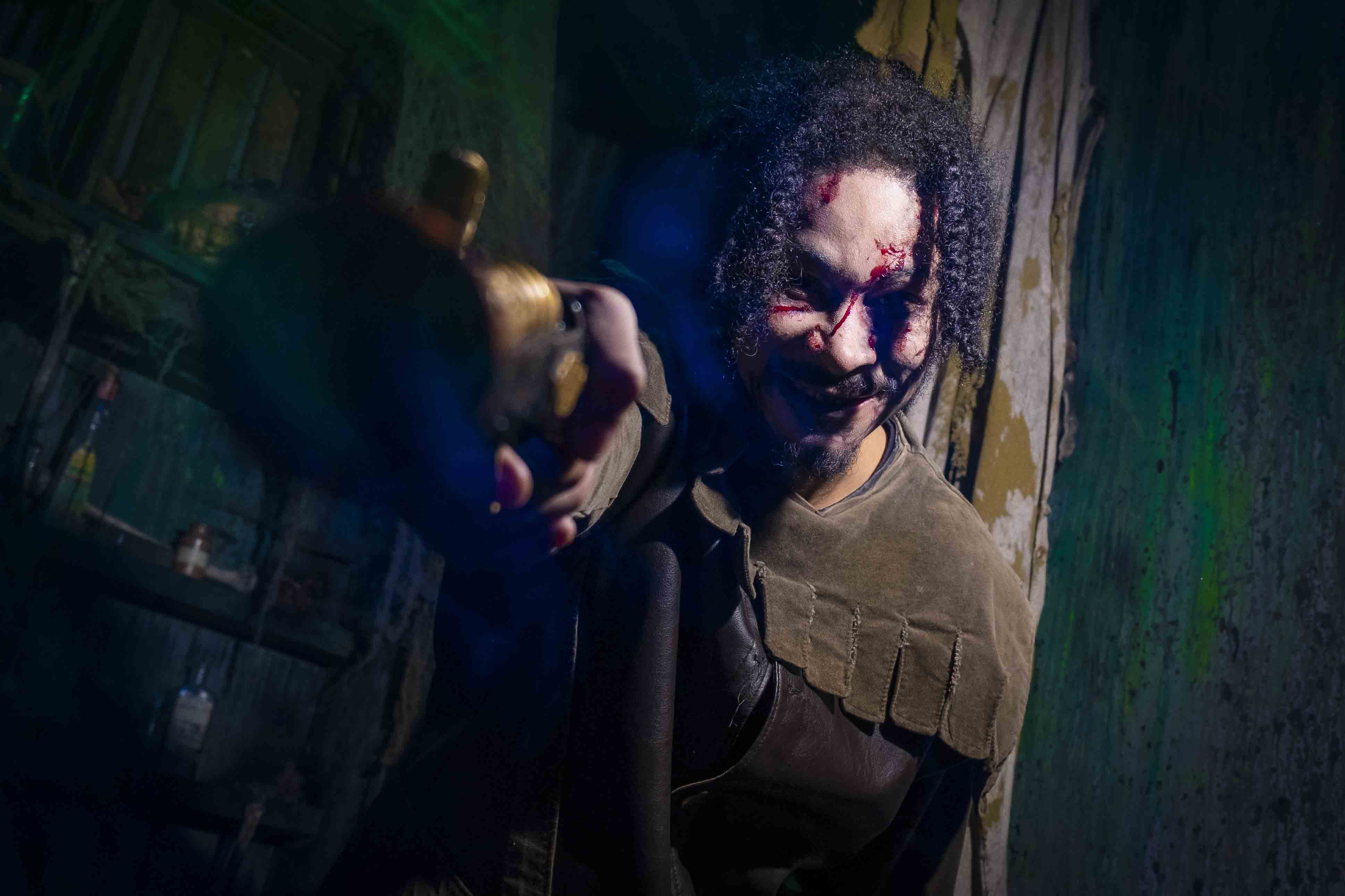 A person with blood on their face stands in a dimly lit, grungy room, pointing an object towards the camera, evoking a tense, mysterious atmosphere.