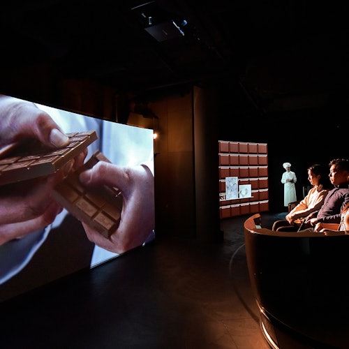 People seated in a dark room watching a large screen showing hands breaking a chocolate bar; an illuminated mannequin stands nearby.
