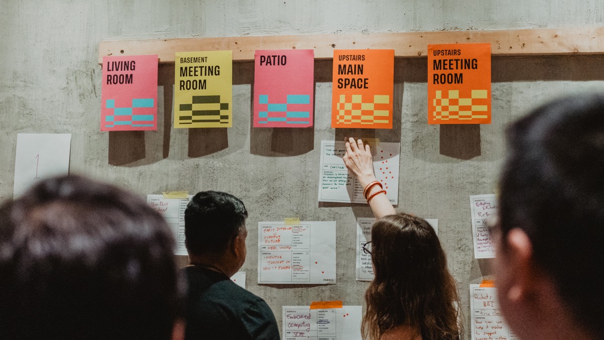 Two people stand in front of a wall with colorful signs labeled "Living Room," "Basement Meeting Room," "Patio," "Upstairs Main Space," and "Upstairs Meeting Room." They are discussing and arranging papers.