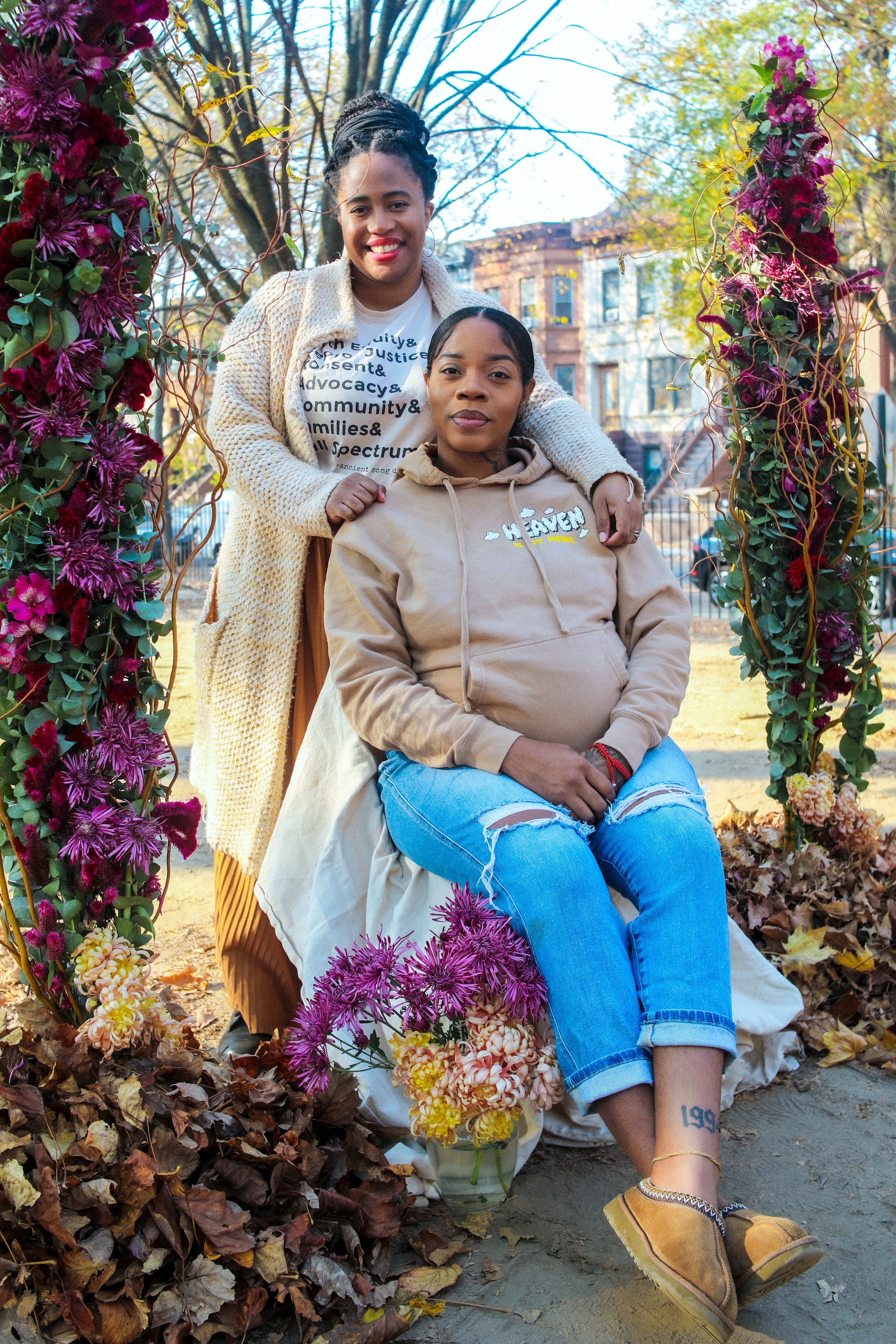 A woman sits gracefully among blooming flowers and autumn leaves, wearing a light brown hoodie and jeans, while another stands beside her in a cream cardigan, both in an urban park with historic buildings in the background.
