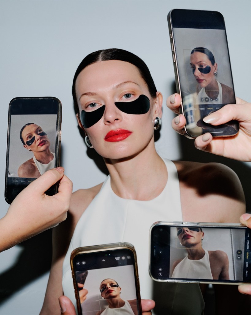 A woman with black eye masks poses for photos while being surrounded by multiple smartphones capturing her image.