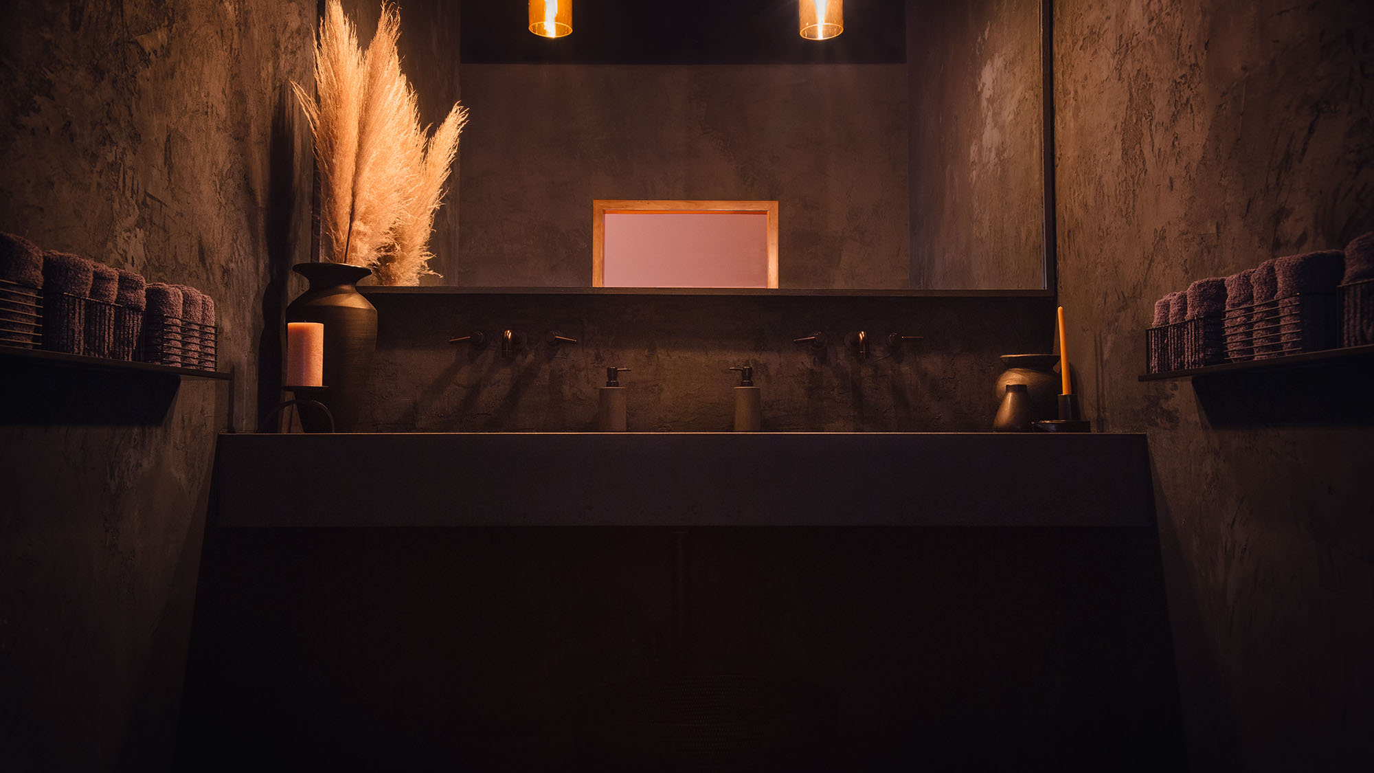 A concrete sink is hung between two concrete walls adourned wth eco-friendly towels. The sink is decorated with a yellow dried plant resembling wheat and candles of the same hue. The mirror reveals a doorway behind camera which is illuminated by the sunset in a wash of pink color. This is the washroom sink at Particle Studios in Seattle, WA.