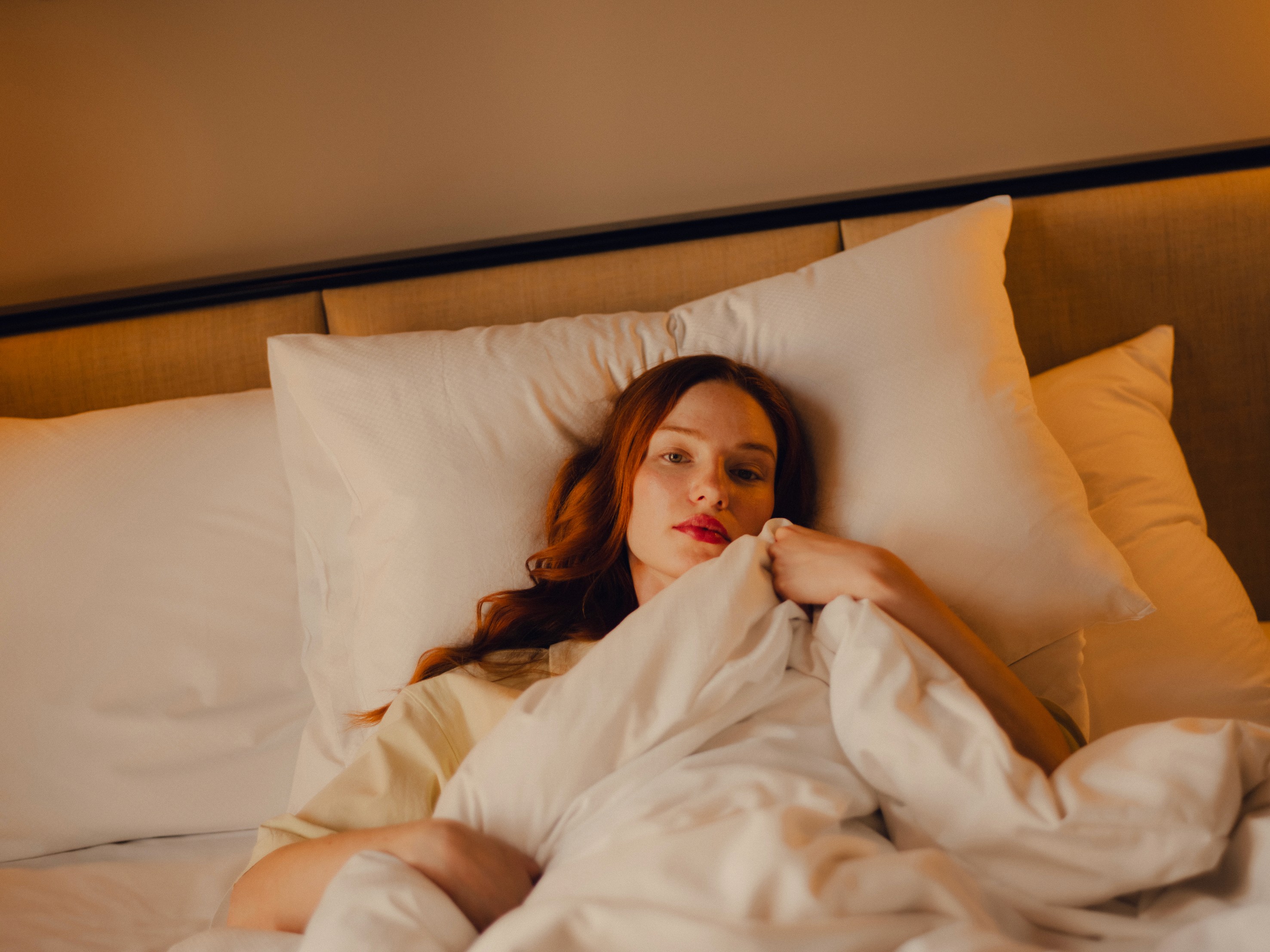 A young woman with red hair lying in bed, propped up by white pillows and wrapped in a white duvet, her expression relaxed yet pensive under warm ambient lighting.