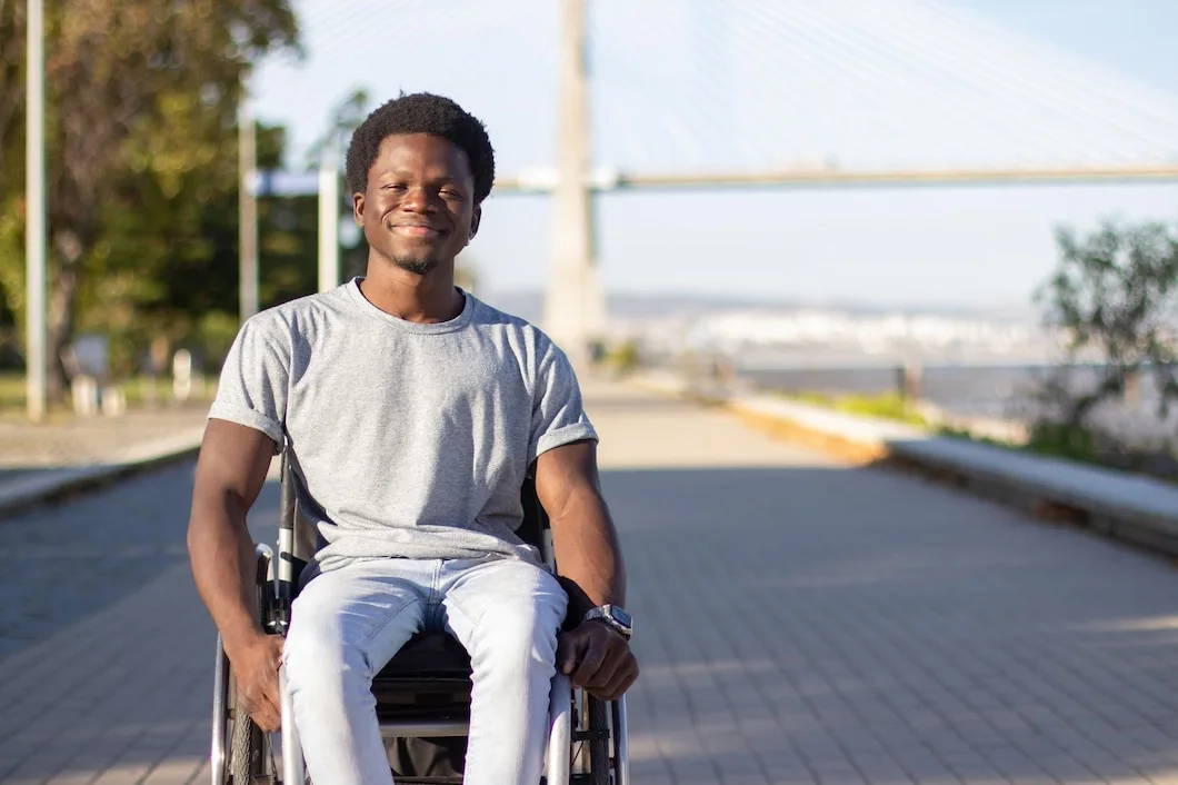 Homem jovem, negro, sorrindo e sentado em uma cadeira de rodas ao ar livre. Ele usa uma camiseta cinza e calça jeans clara. Ao fundo, desfocado, é possível ver uma ponte moderna e algumas árvores, sugerindo que ele está em um parque ou calçadão em um dia ensolarado.