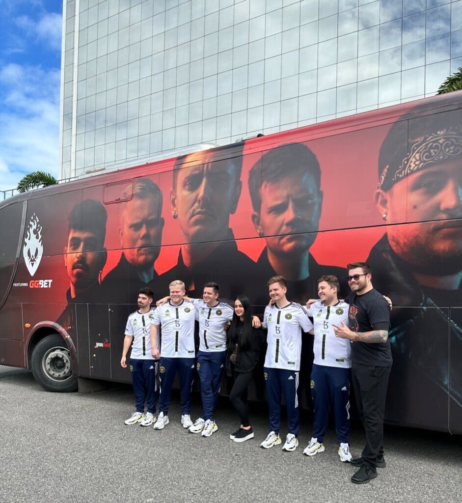 Visuel du projet Metality : Équipe Metality accompagnée d'une femme et d'un homme habillés en noir, se tenant devant leur bus de tournée rouge et noir arborant les visages de l'équipe.