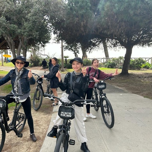 Cuatro personas de pie con bicicletas en un sendero pavimentado en un parque, sonriendo y haciendo señales de aprobación con el pulgar. Hay árboles y vegetación en el fondo.