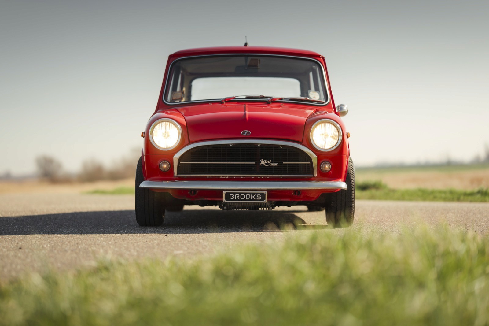 A bright red vintage car parked on a rural road, surrounded by green grass and an open sky.