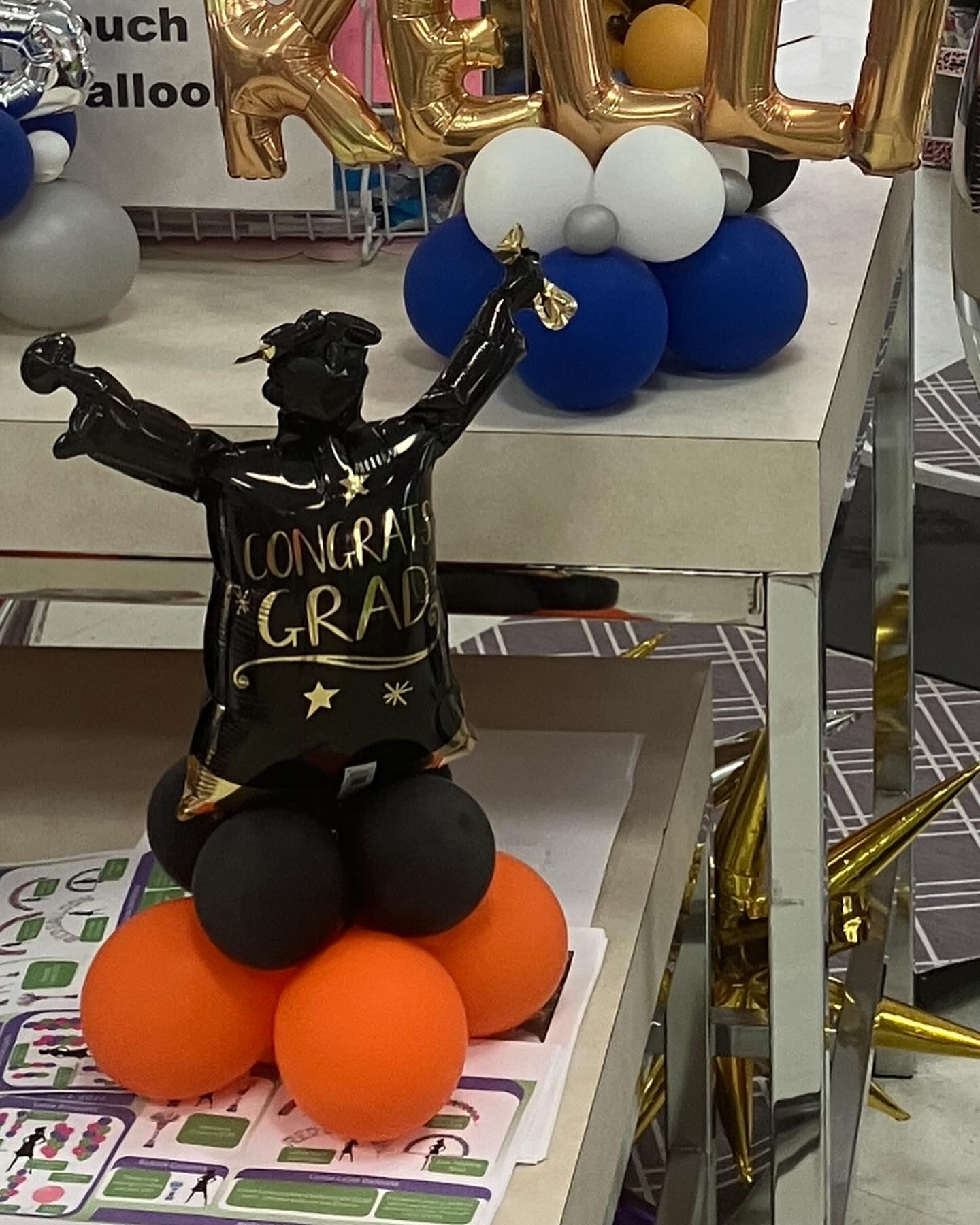 Black and gold graduation balloon figure with raised arms reading Congrats Grad displayed on a base of black and orange balloons created by Be Bold Balloons