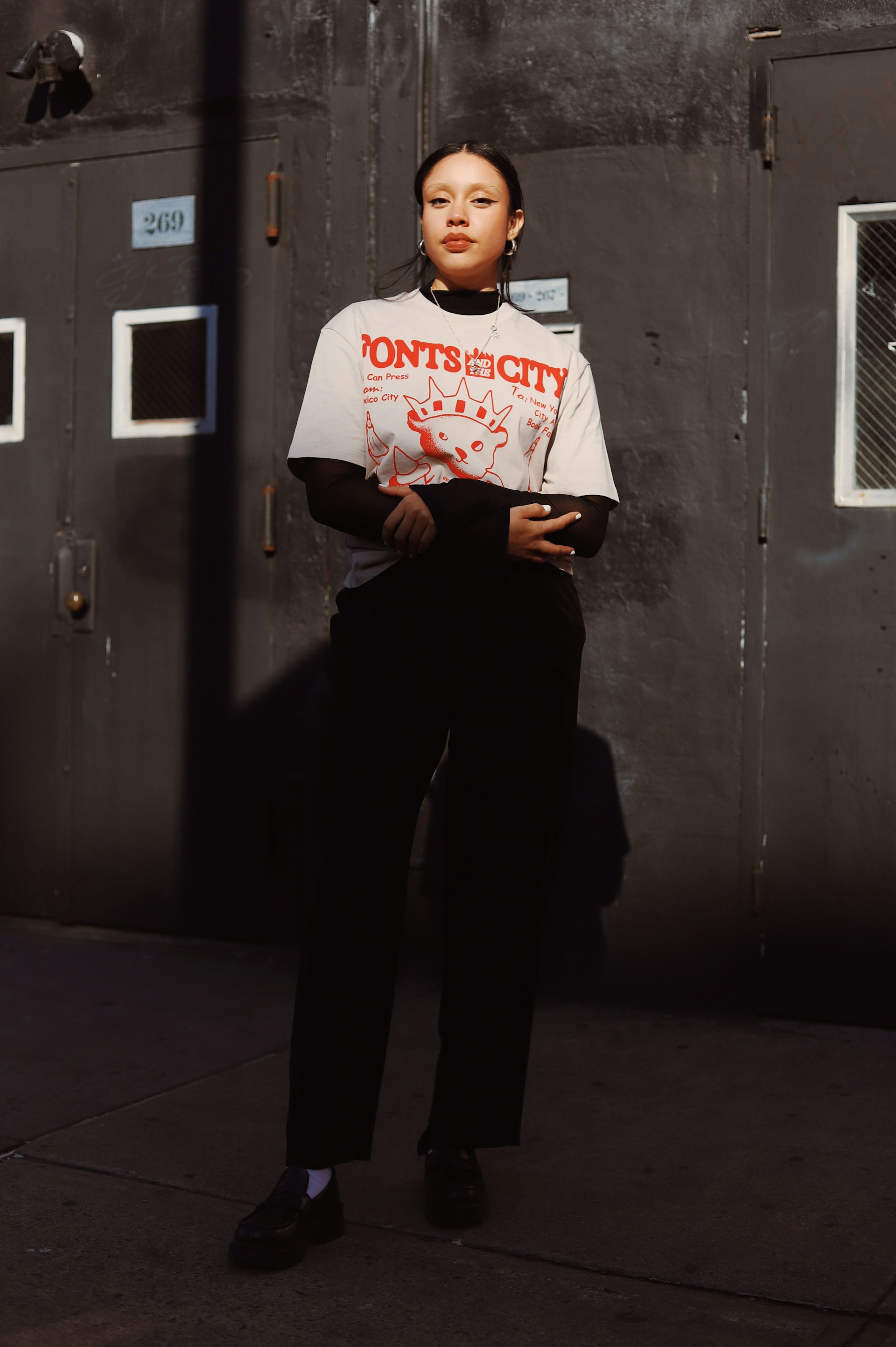A full-length photo of the woman posing in front of a dark building. She is dressed in a grey t-shirt, black mesh top, black pants, and loafers, looking toward the camera with a neutral expression.