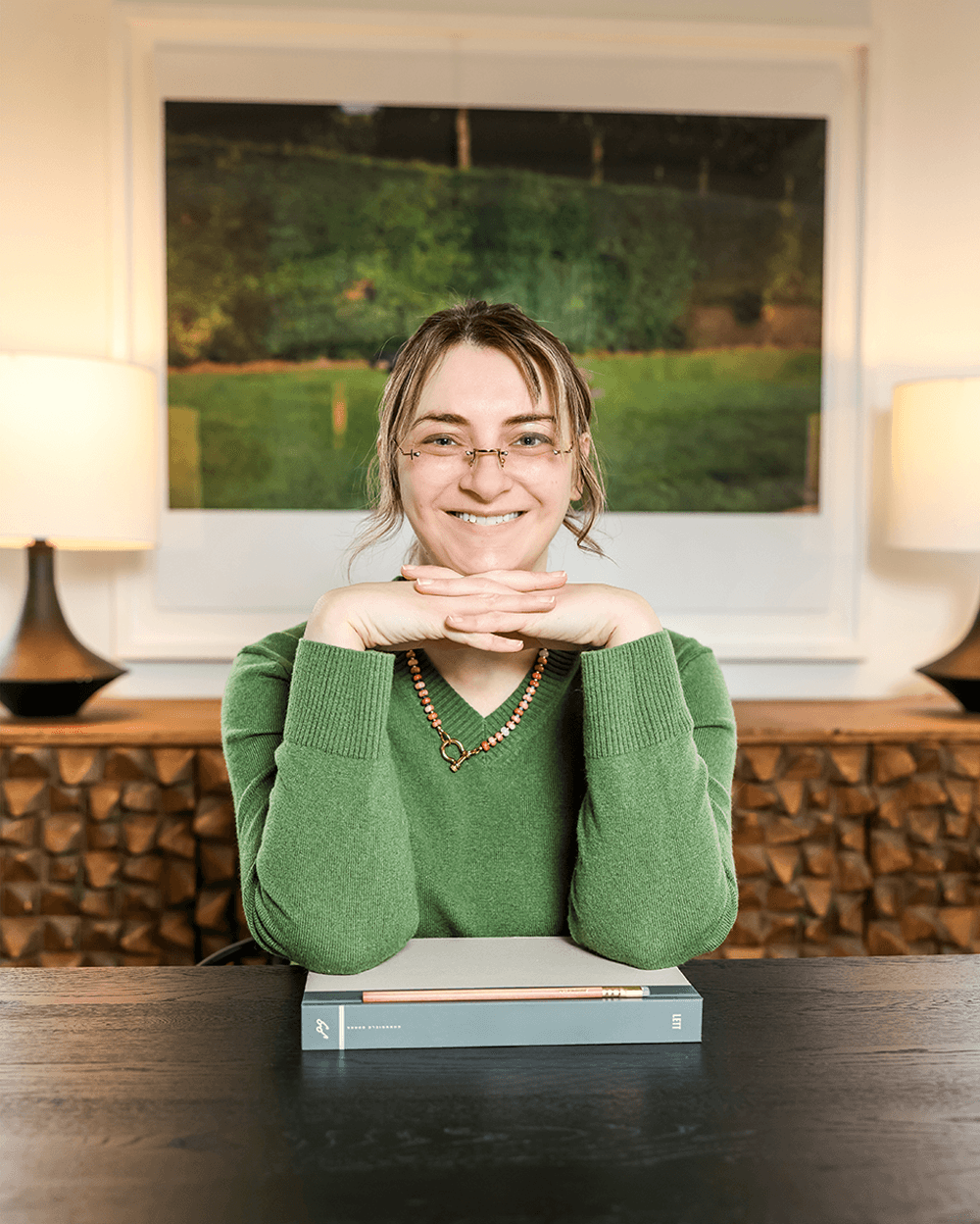 A smiling woman with shoulder-length blonde hair and round glasses sits outdoors wearing a sage green eyelet lace blouse with intricate cutout patterns and three-quarter sleeves, paired with a navy skirt and delicate necklace. She's positioned against a natural backdrop of lush green trees and grass on what appears to be a sunny day.