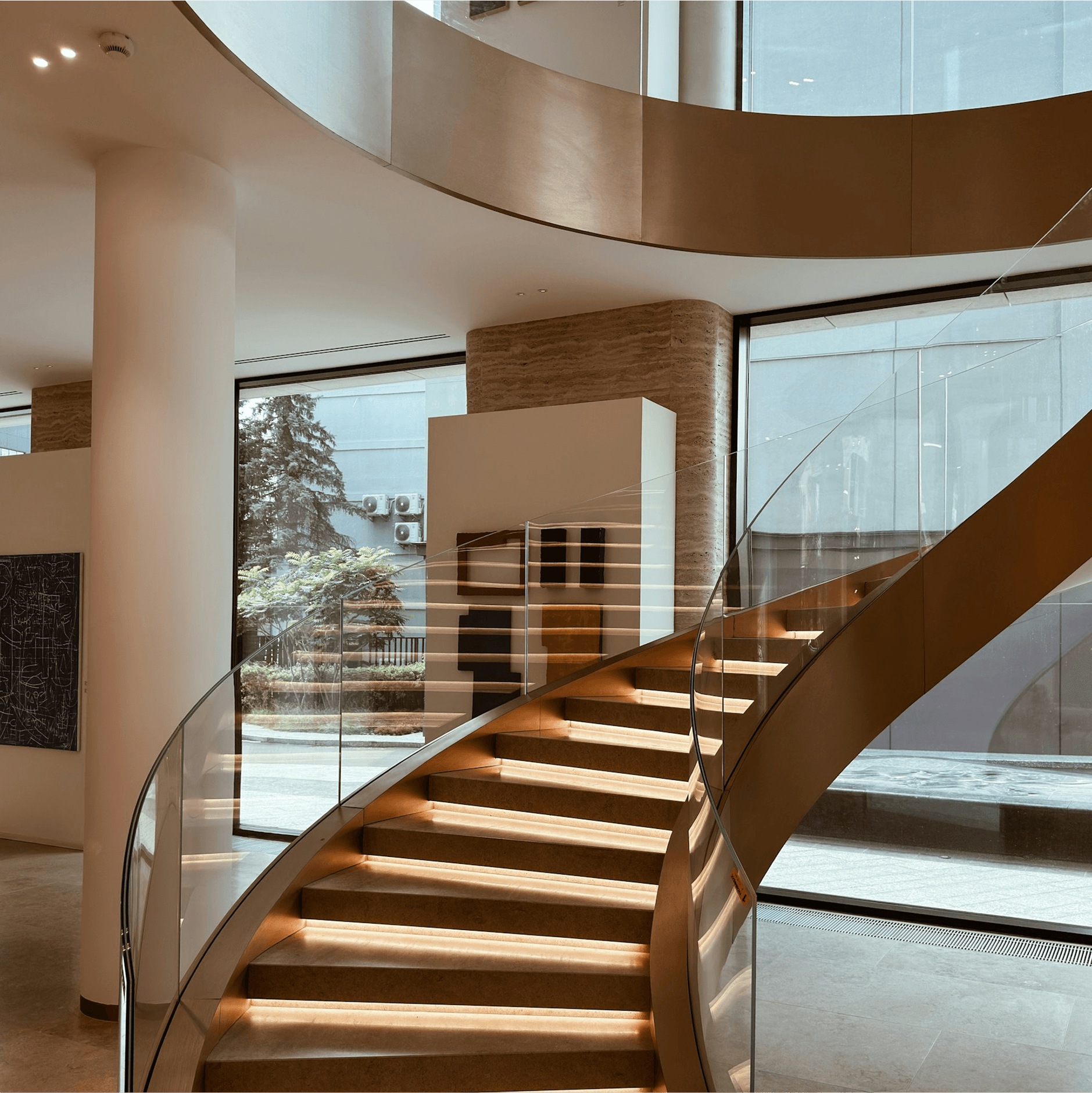 A modern, spiral staircase with wooden steps and glass railings is elegantly illuminated, leading to an upper floor in a contemporary gallery space, surrounded by large windows allowing natural light to flood in.