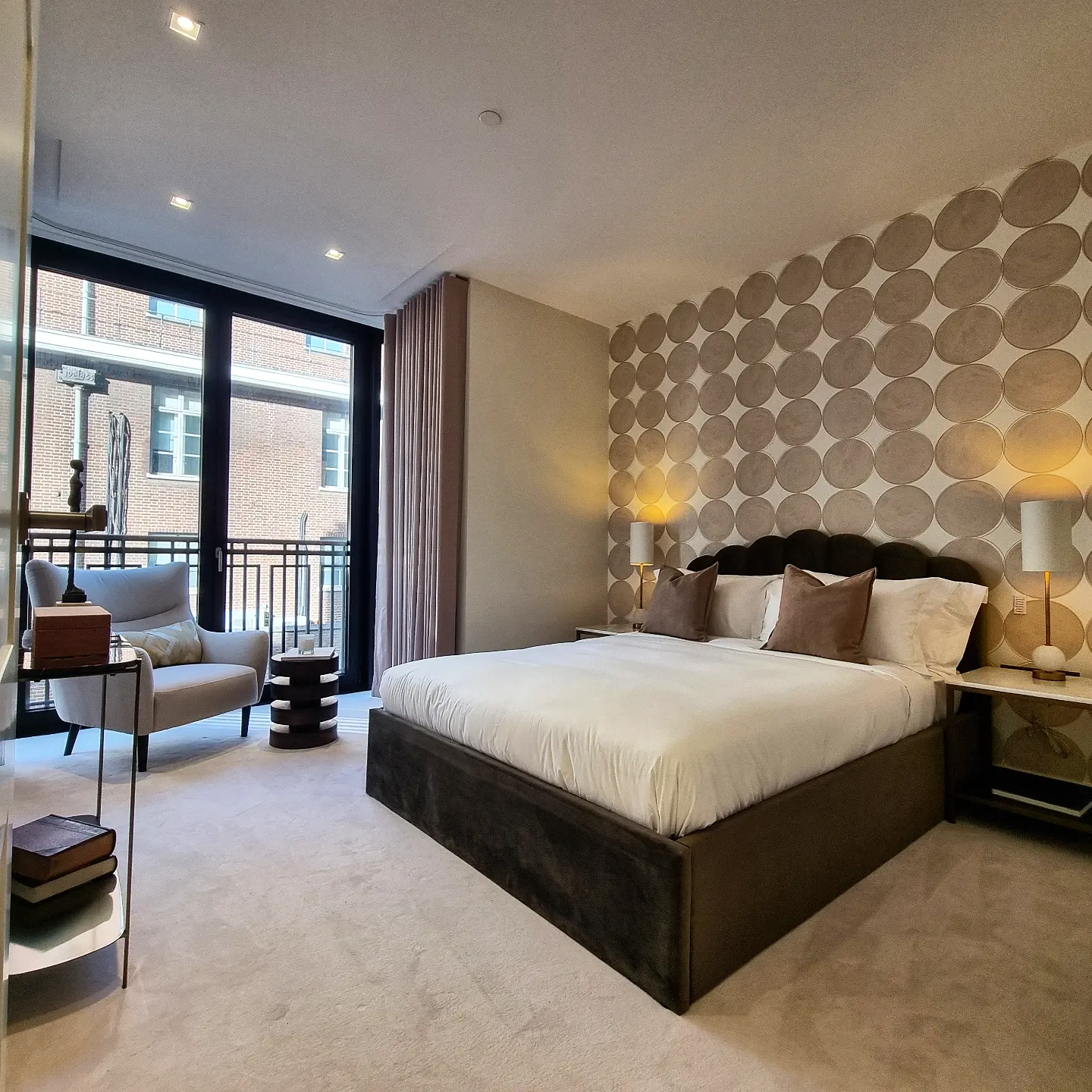 Luxurious modern bedroom featuring a large bed with white linens and brown accent pillows, flanked by sleek bedside lamps, complemented by a textured accent wall with circular patterns and a cozy seating area by floor-to-ceiling windows overlooking an urban scene.