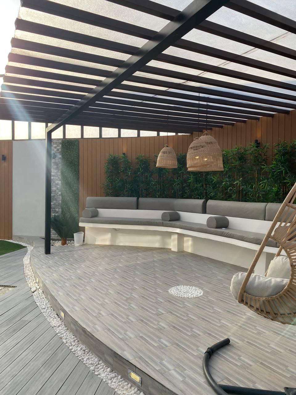 A modern outdoor patio with seating under a slatted pergola, surrounded by greenery and paved flooring.