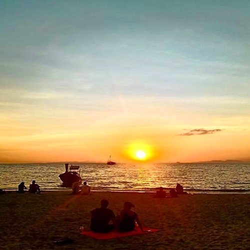 Mga tao na nakaupo sa dalampasigan habang papalubog ang araw, nanonood ng mga bangka sa dagat. Ang kalangitan ay kulay kahel at asul.