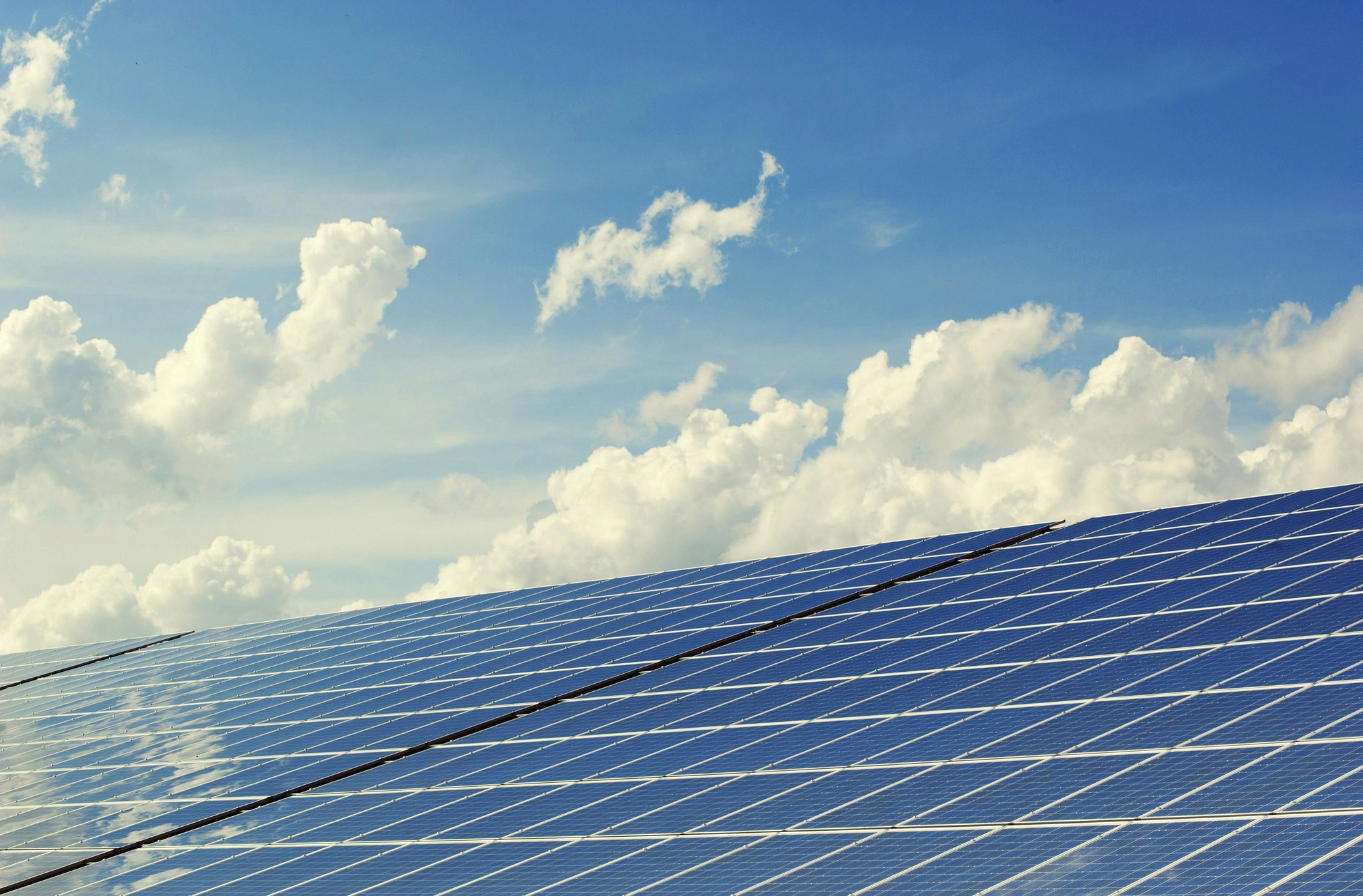  A large array of solar panels under a clear blue sky with fluffy white clouds.