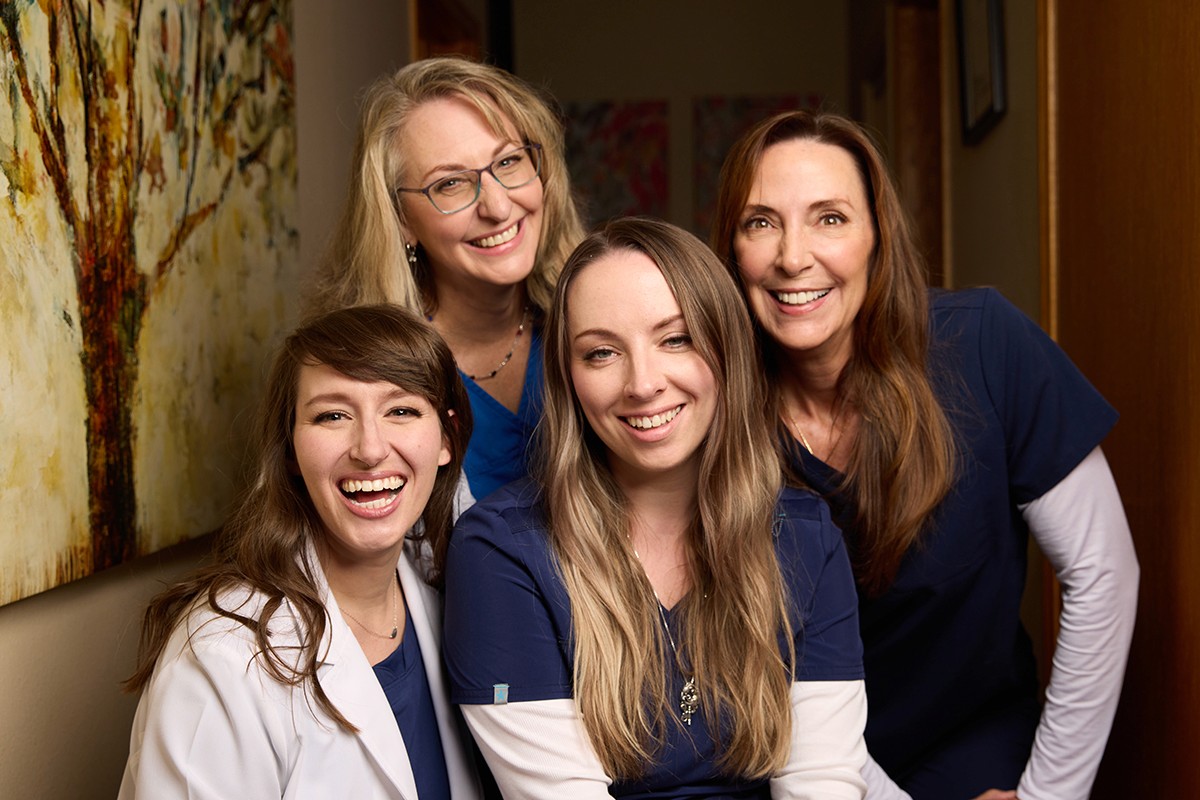 Four women in audiology uniforms smiling together in a warm, friendly setting.