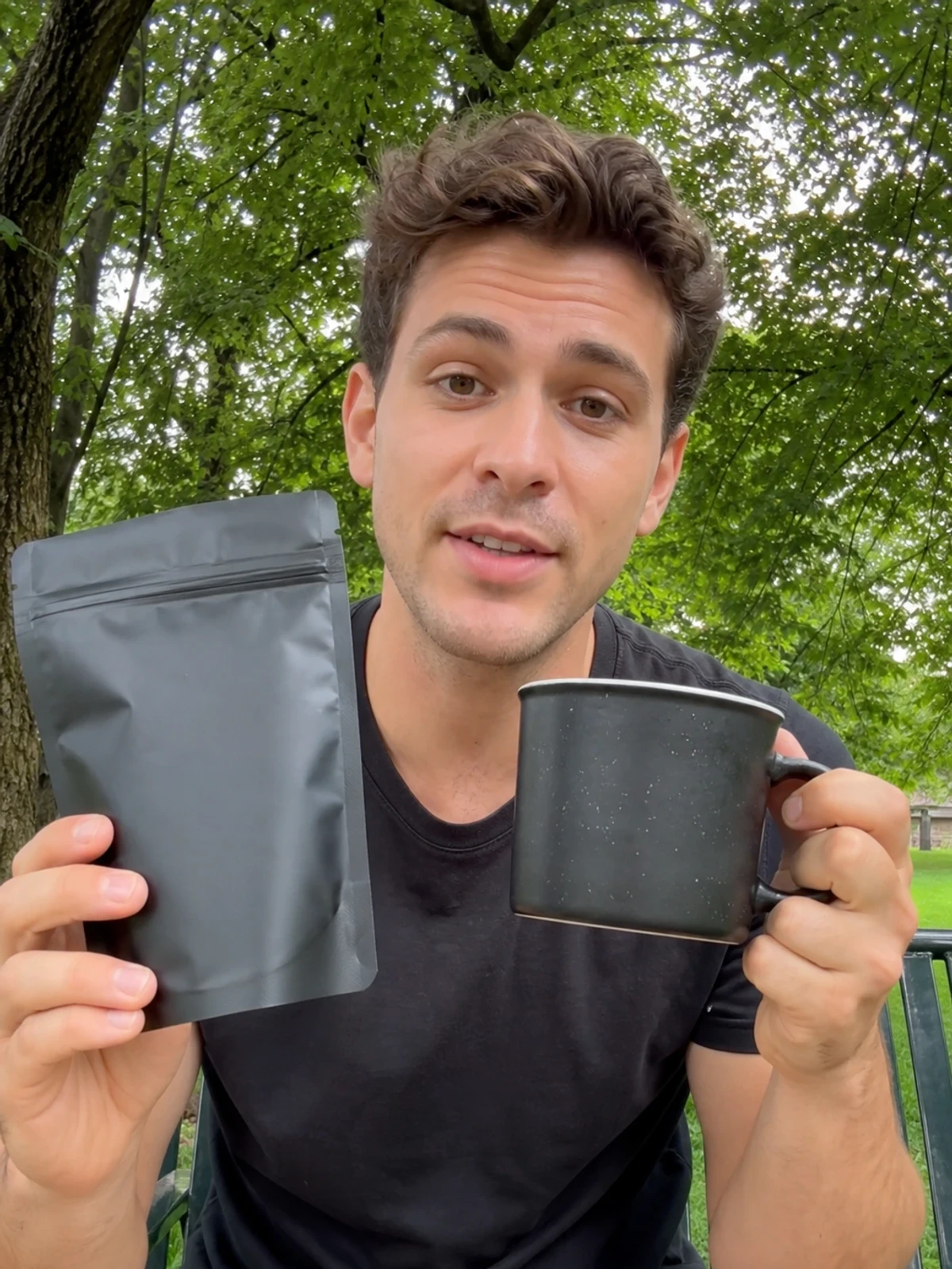 UGC-style photo: young male creator, on a park bench, showing a coffee pouch and mug while talking to camera, close vertical talking-head shot.