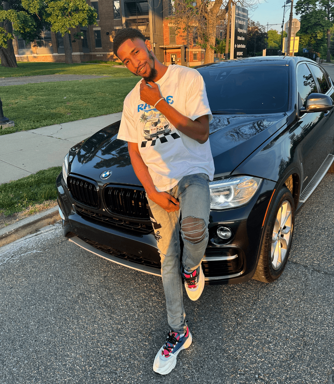 Tyrone “TyFix” Adeyeye leans against a black BMW SUV during golden hour, wearing casual streetwear.
