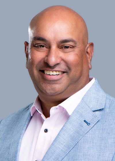 Blonde woman in a black long-sleeve and apron, arms crossed—minimalist studio portrait on a beige background.
