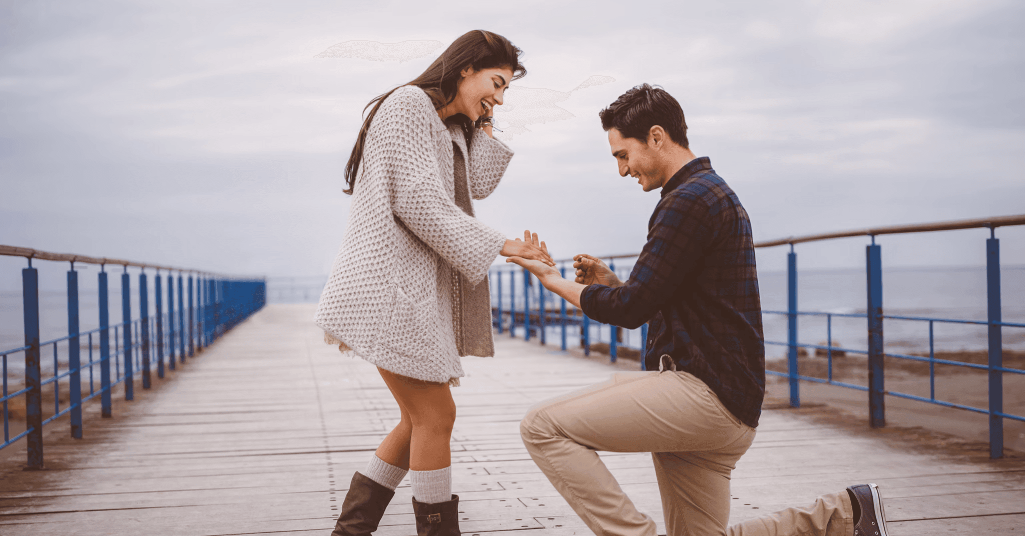 En man på ett knä på en hamnbrygga friar till en kvinna genom att sätta en ring på hennes finger, medan hon, överväldigad av känslor, täcker sin mun med handen i ett uttryck av förvåning och glädje.