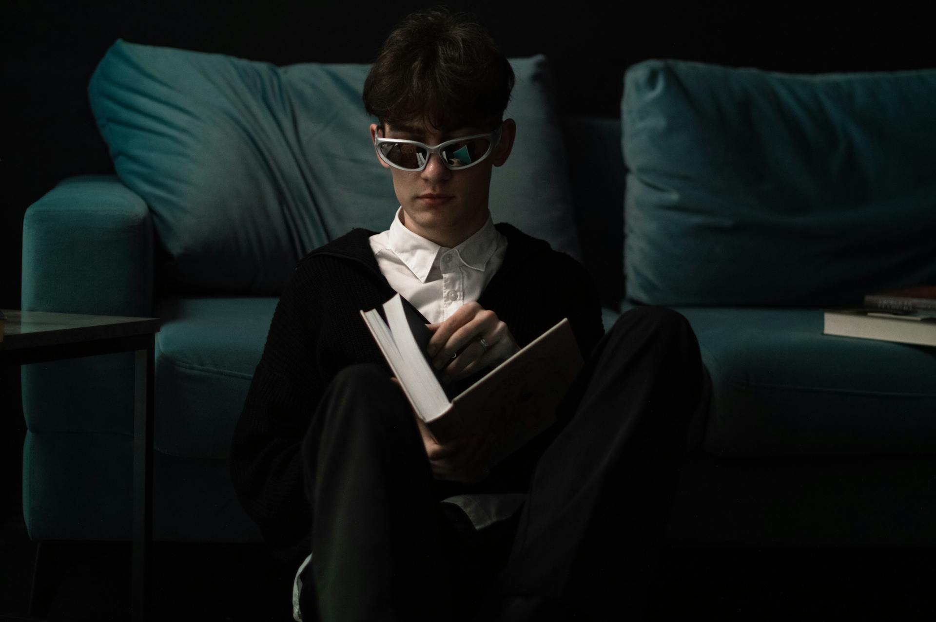 A young person in silver sports sunglasses, a white shirt, and a black sweater sits on the floor, holding an open book and leaning against a teal couch.