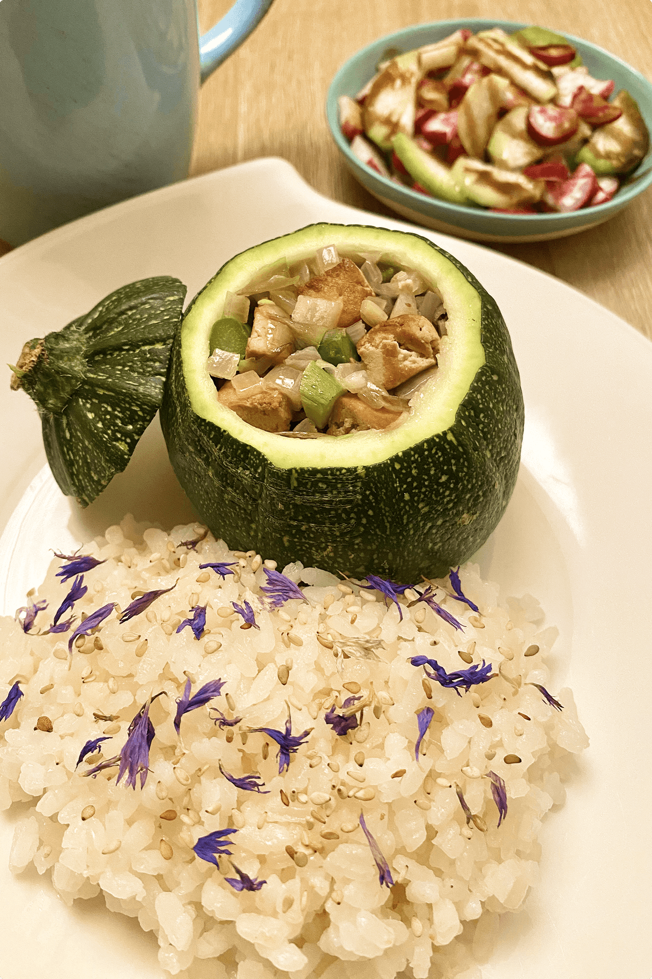 Courgette ronde farcie de poulet et légumes, servie avec du riz aux graines de sésame et pétales de bleuet. Une salade est en arrière-plan.