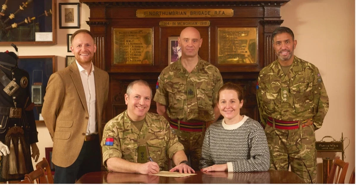 A group of people, including several in military uniforms and one in civilian clothing, pose indoors around a wooden table where a document is being signed. Behind them is a wooden memorial display with engraved plaques and framed items.