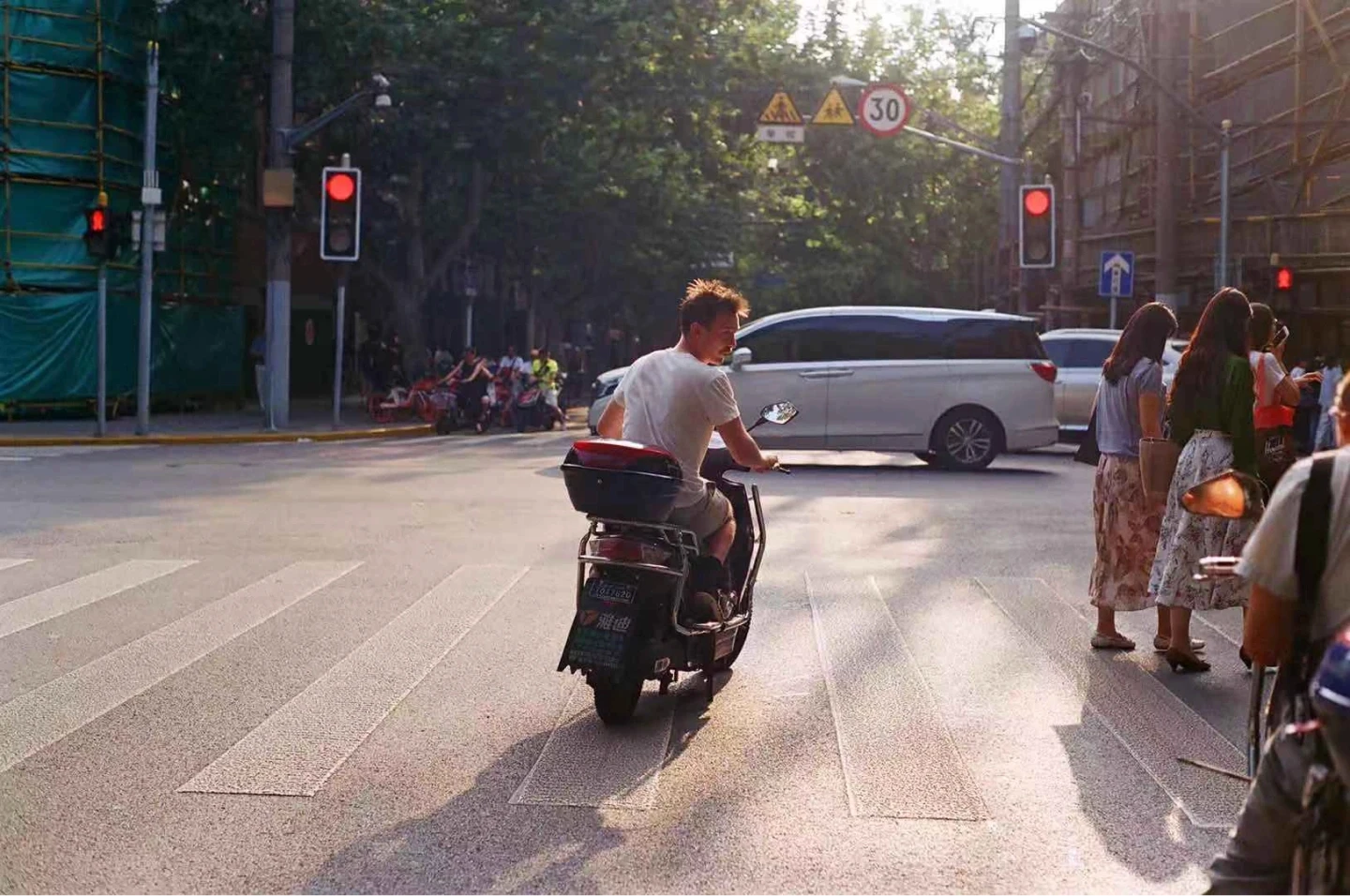 A person on a scooter rides through a sunlit urban intersection with a van approaching and pedestrians waiting at the crosswalk, surrounded by traffic lights and tree-lined streets.