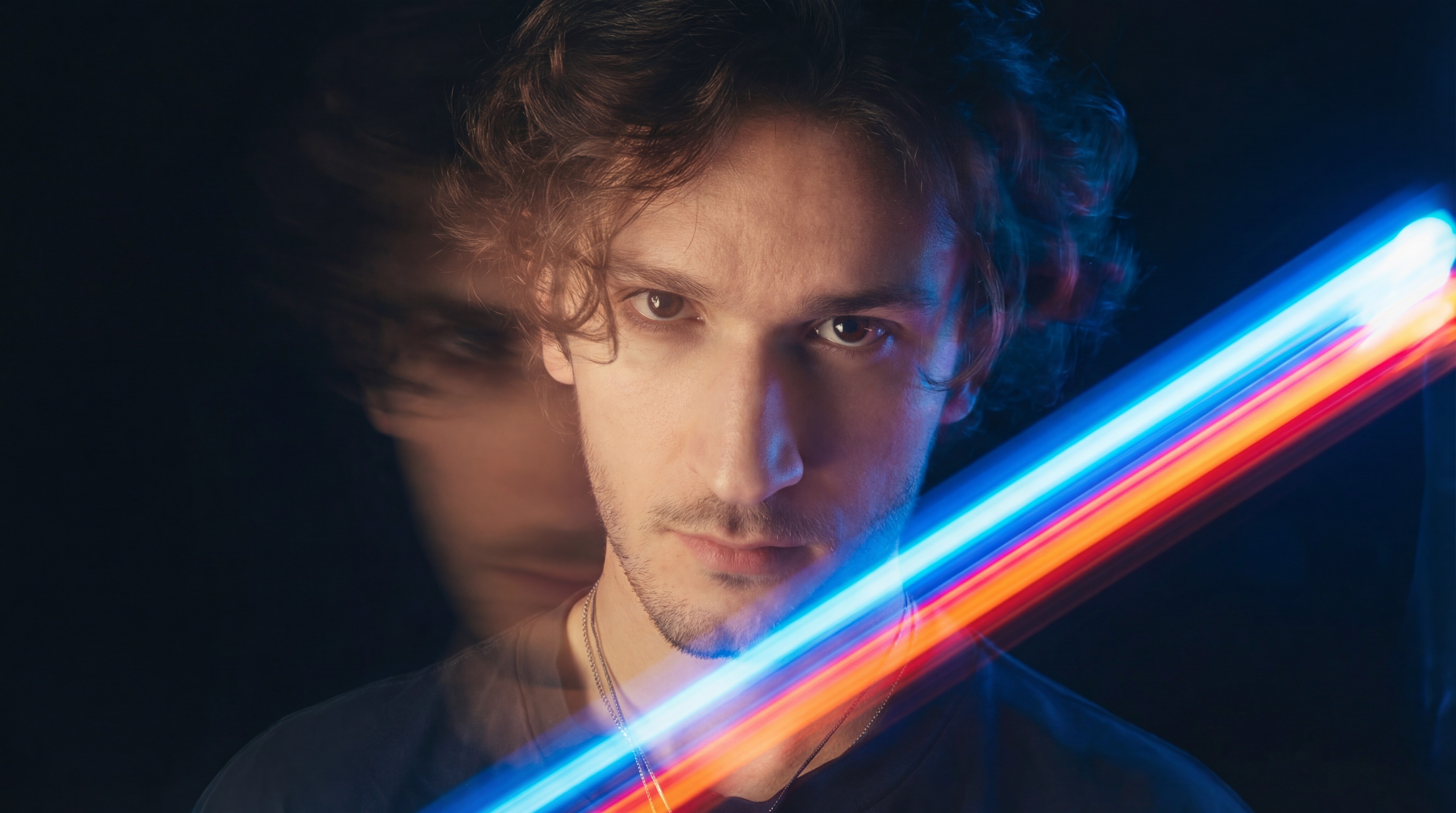 Artistic portrait of young man with curly hair in black shirt and chain, multiple exposure with orange-golden light and neon blue streaks, dynamic motion blur on black background