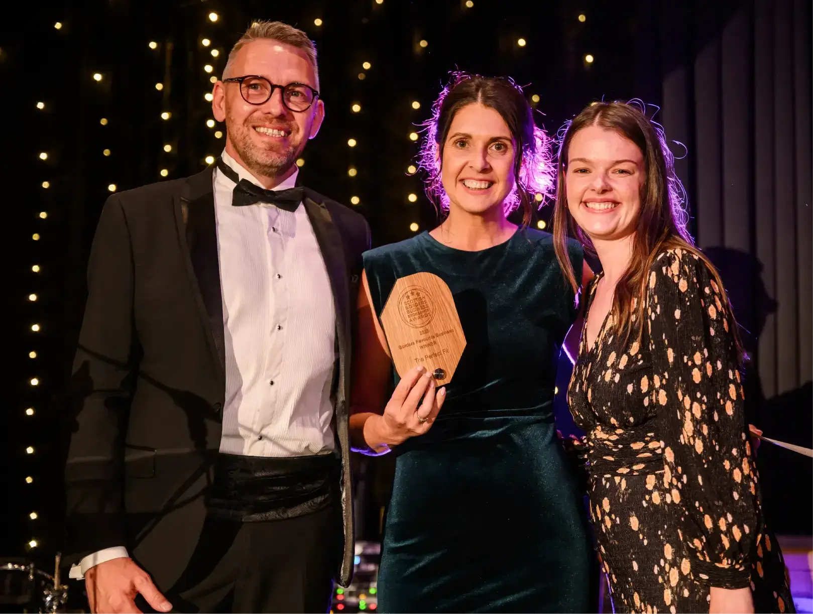 Three people smiling at a formal event. The woman in the center holds a wooden award. They stand against a backdrop of twinkling lights.