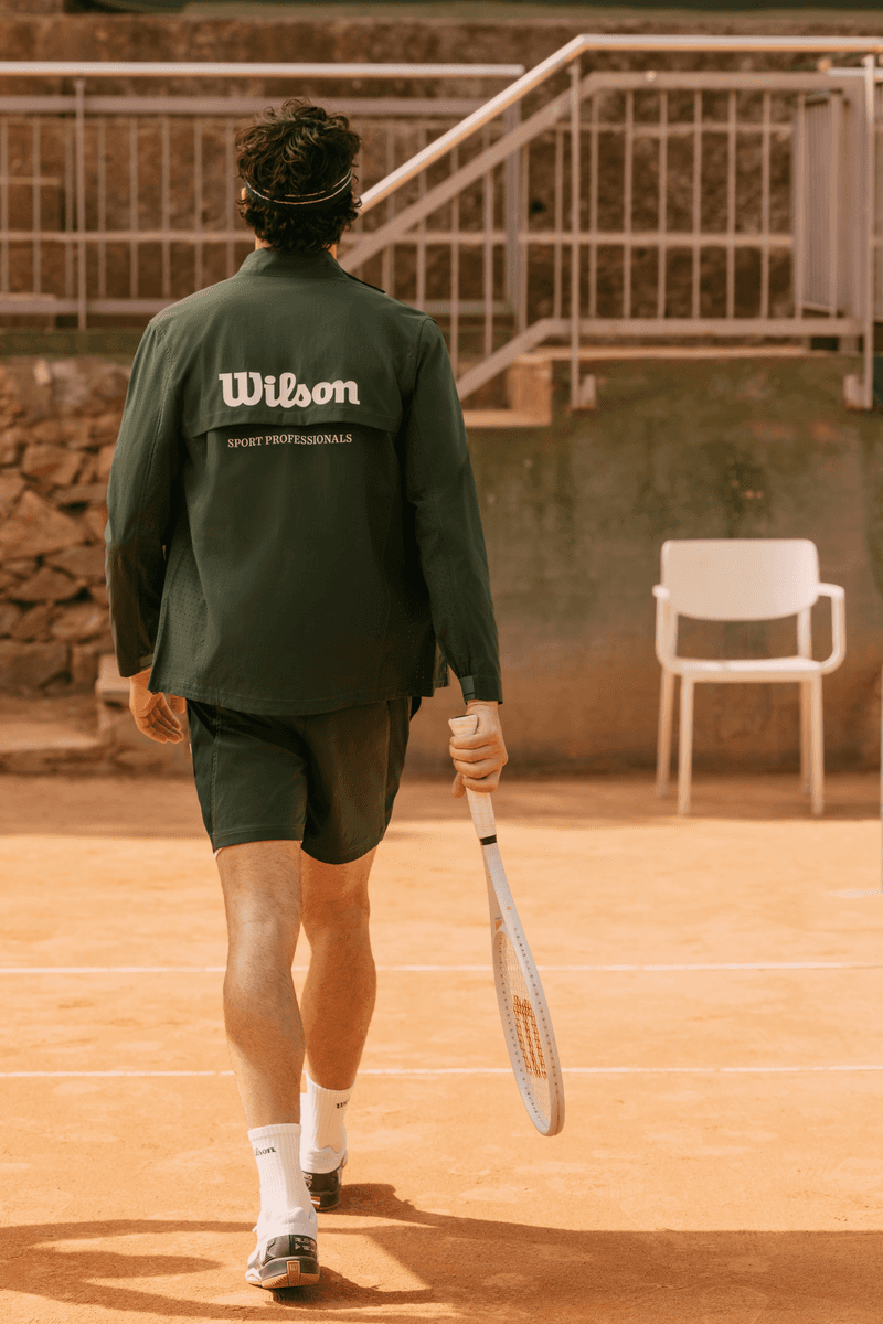 Mannequin vu de dos, portant un blouson technique vert avec lelogo de Wilson Courtstyle, sur un court en terre battue ensoleillé