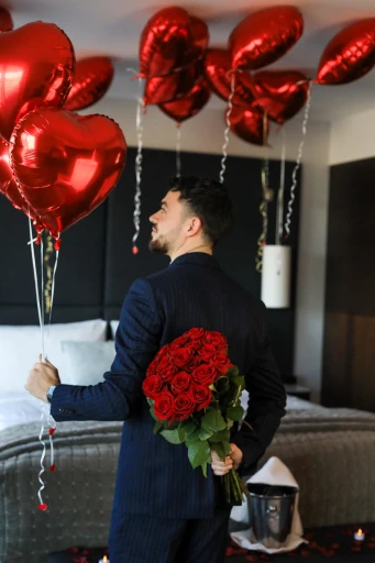 Un homme en costume se tient dans une chambre d'hôtel élégante, tenant un bouquet de roses rouges derrière son dos, entouré de ballons rouges en forme de cœur, évoquant une ambiance romantique.