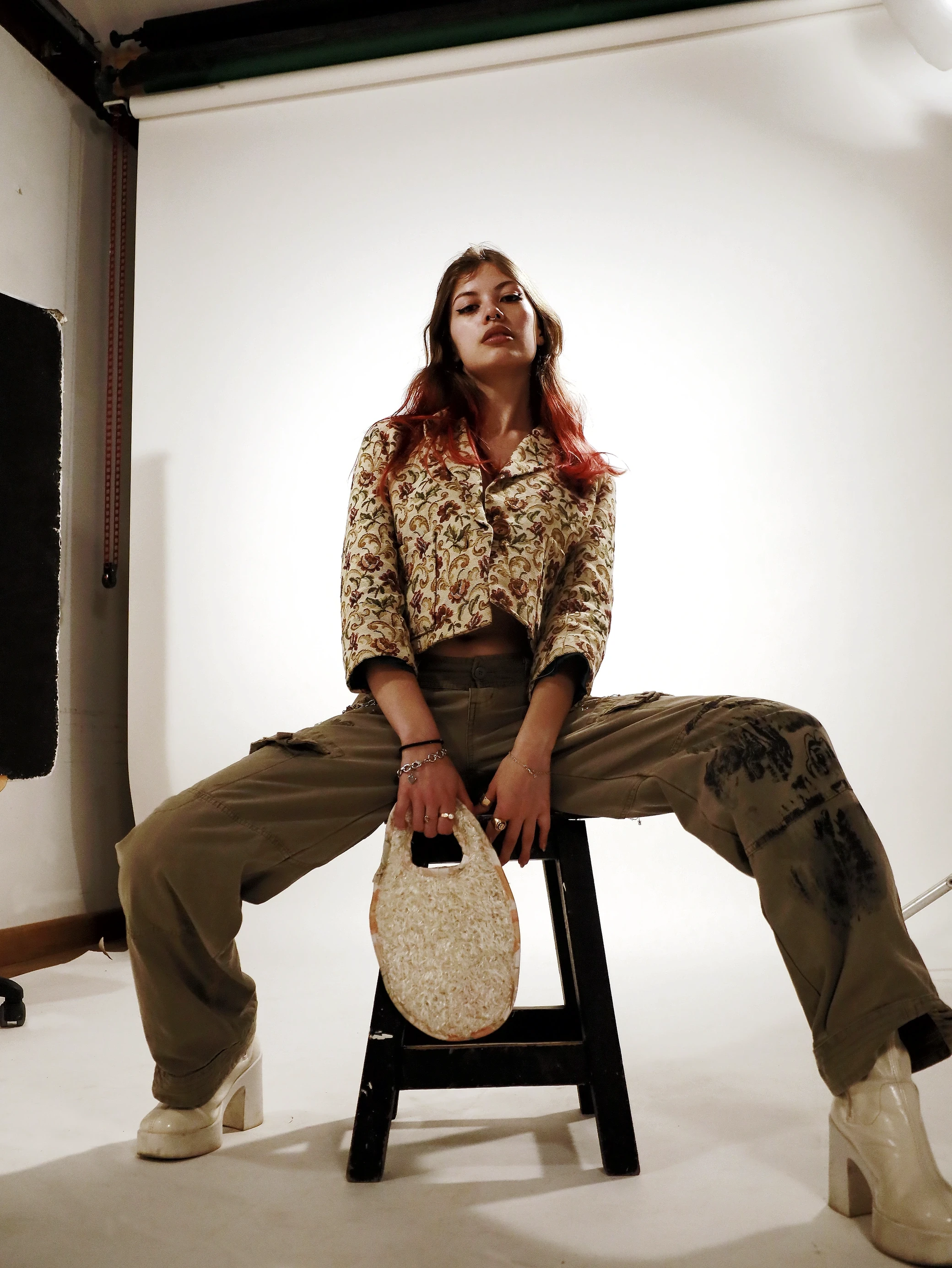 A person in stylish clothing sits on a stool with legs apart, posed against a simple background.