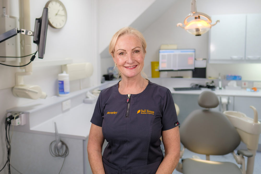 A portrait of Beverley, a Practice Owner at Cricklade Dental Practice, smiling and wearing a dark grey scrub top with a pink zipper pull. The top features yellow embroidered text reading "Beverley" on the left and the practice name on the right. She is standing in a dental surgery with a grey dental chair and medical equipment visible in the background.